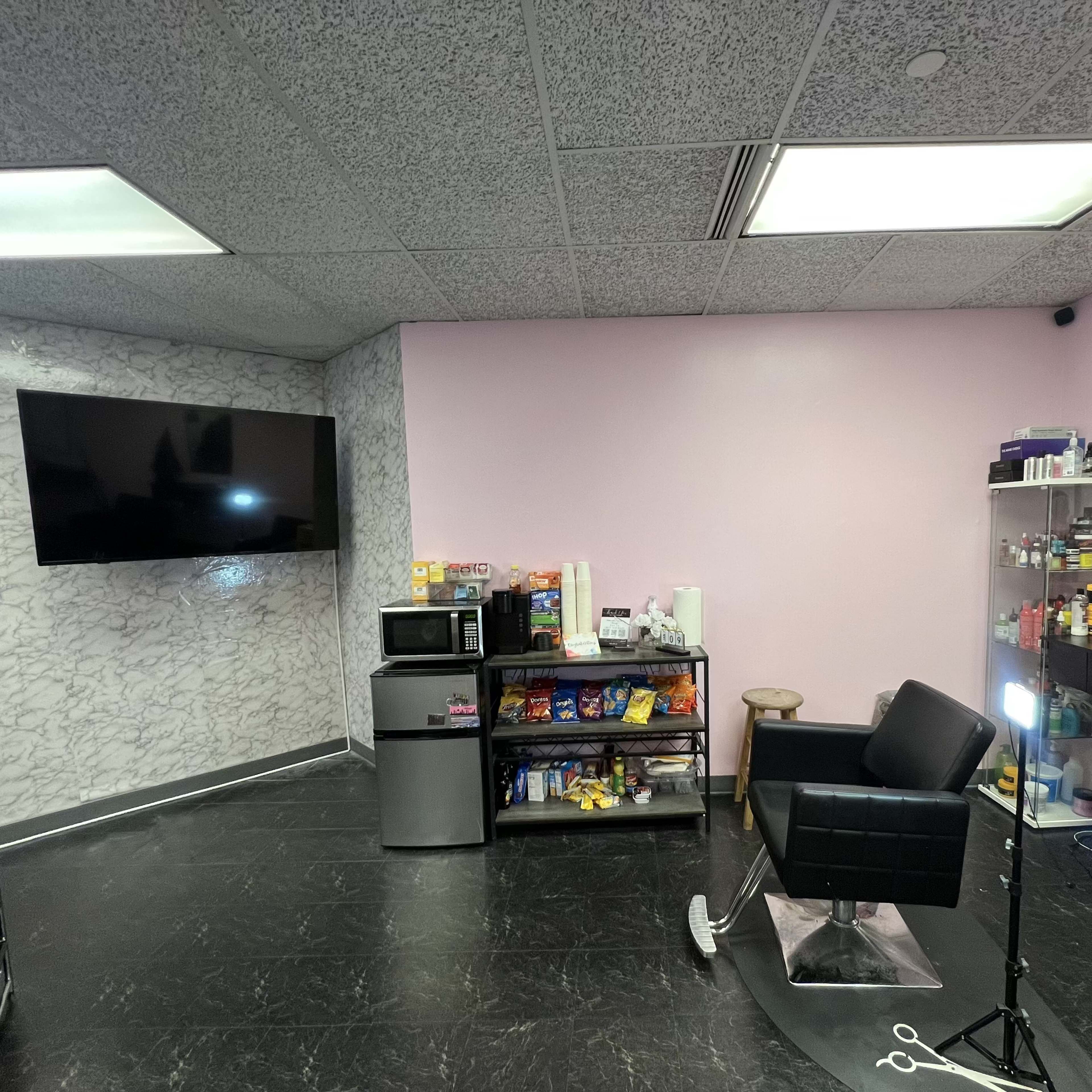 A salon interior with a black barber chair, a television on the wall, a display shelf filled with products, and a small kitchenette area with a microwave and snacks.