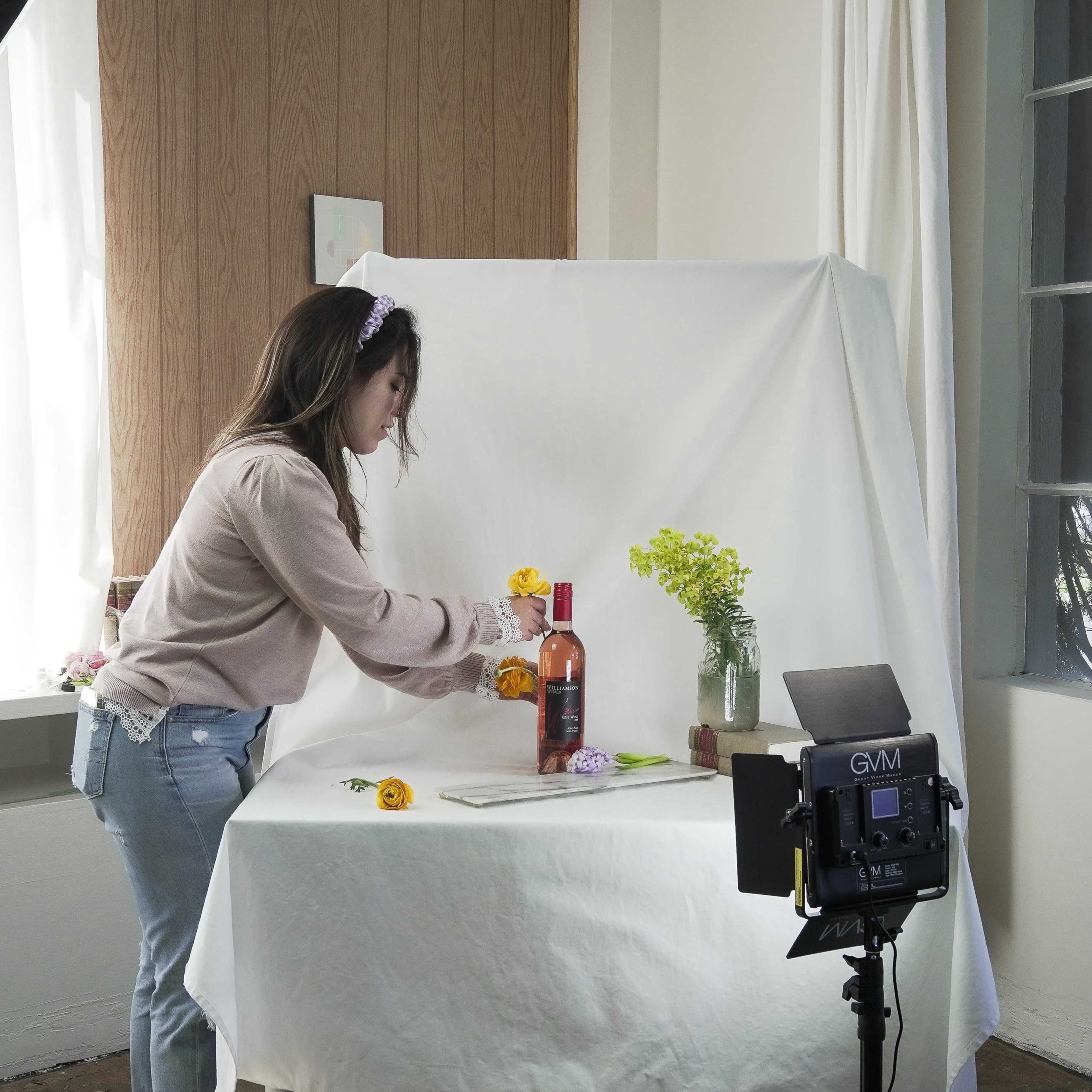 A woman is arranging flowers around a bottle on a table in a bright room set up for photography.