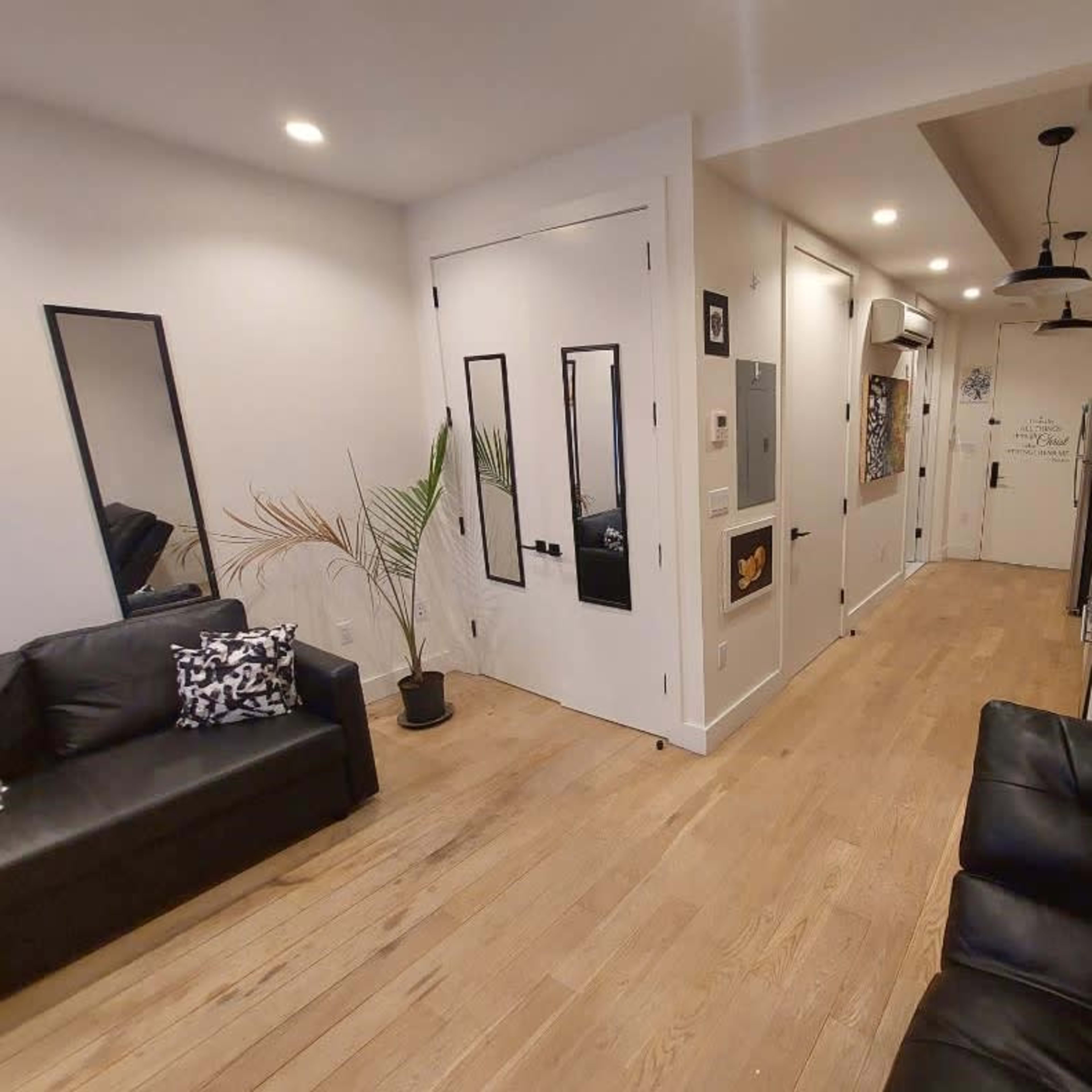 A modern living room with two black sofas, a plant, and a hallway leading to a closed door and various wall decorations.