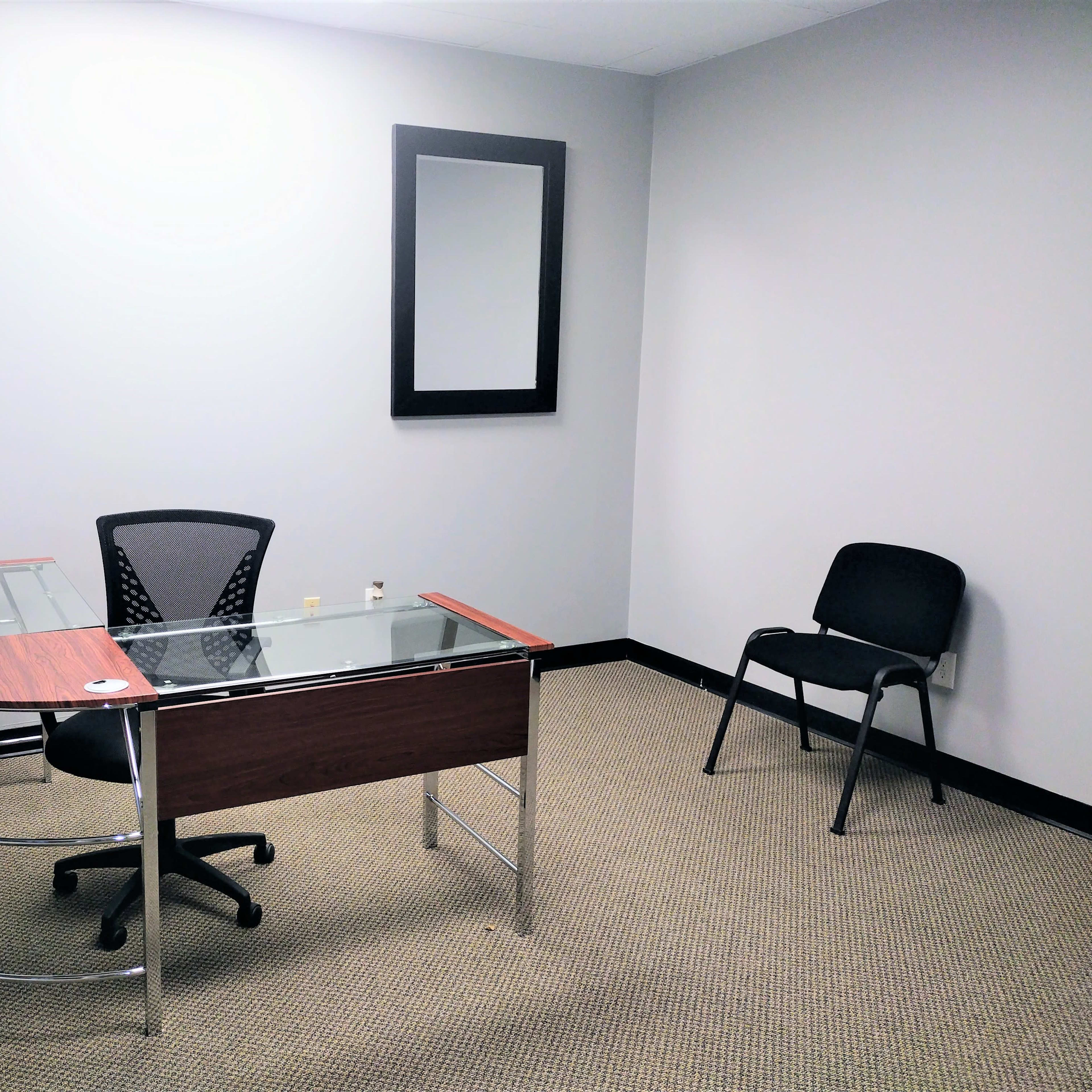 A sparsely furnished office with a glass-top desk, an office chair, a lamp, a mirror on the wall, and a single black chair.