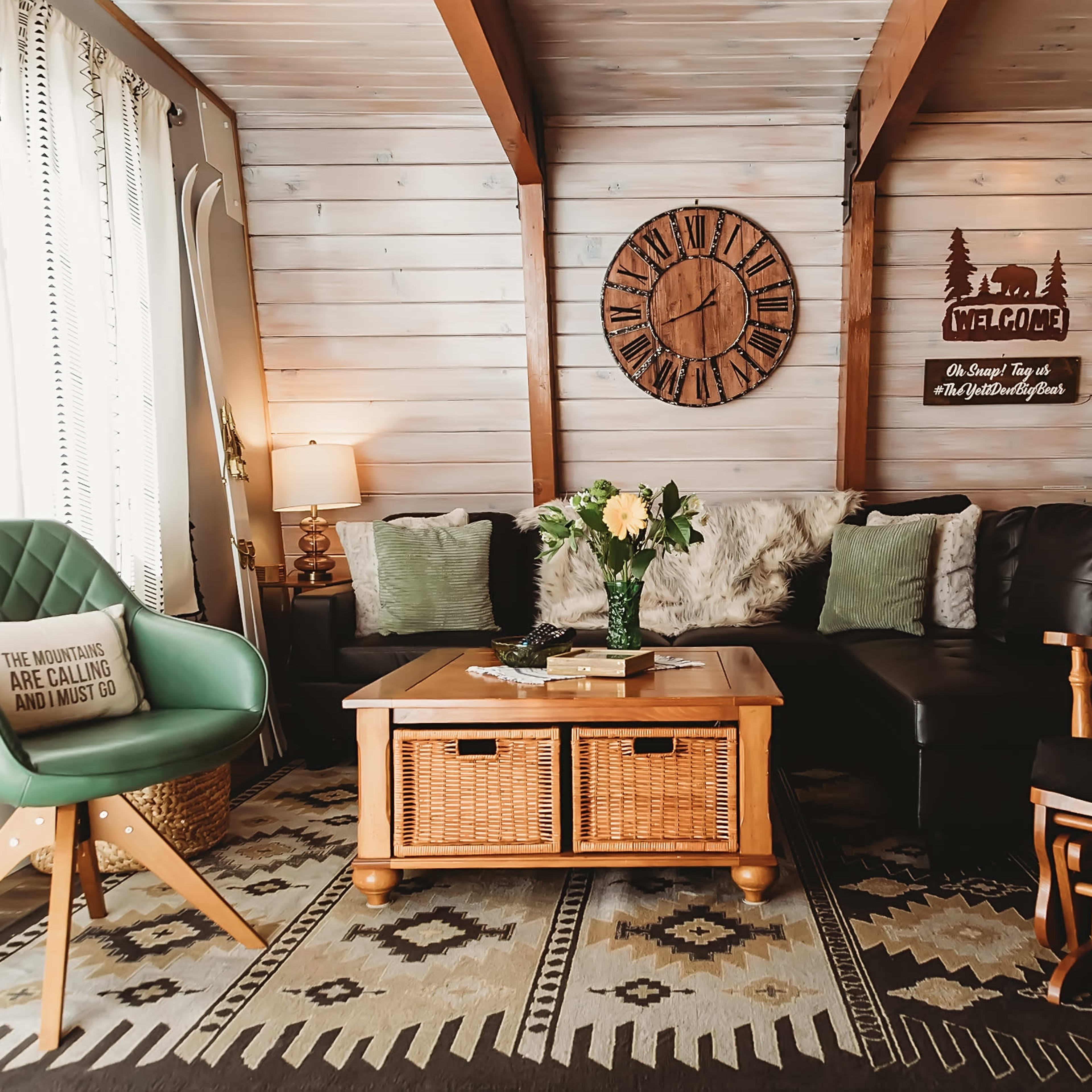 A cozy living room with a wooden ceiling, a clock on the wall, a black couch, a wicker coffee table, and a green chair, decorated with pillows and plants.