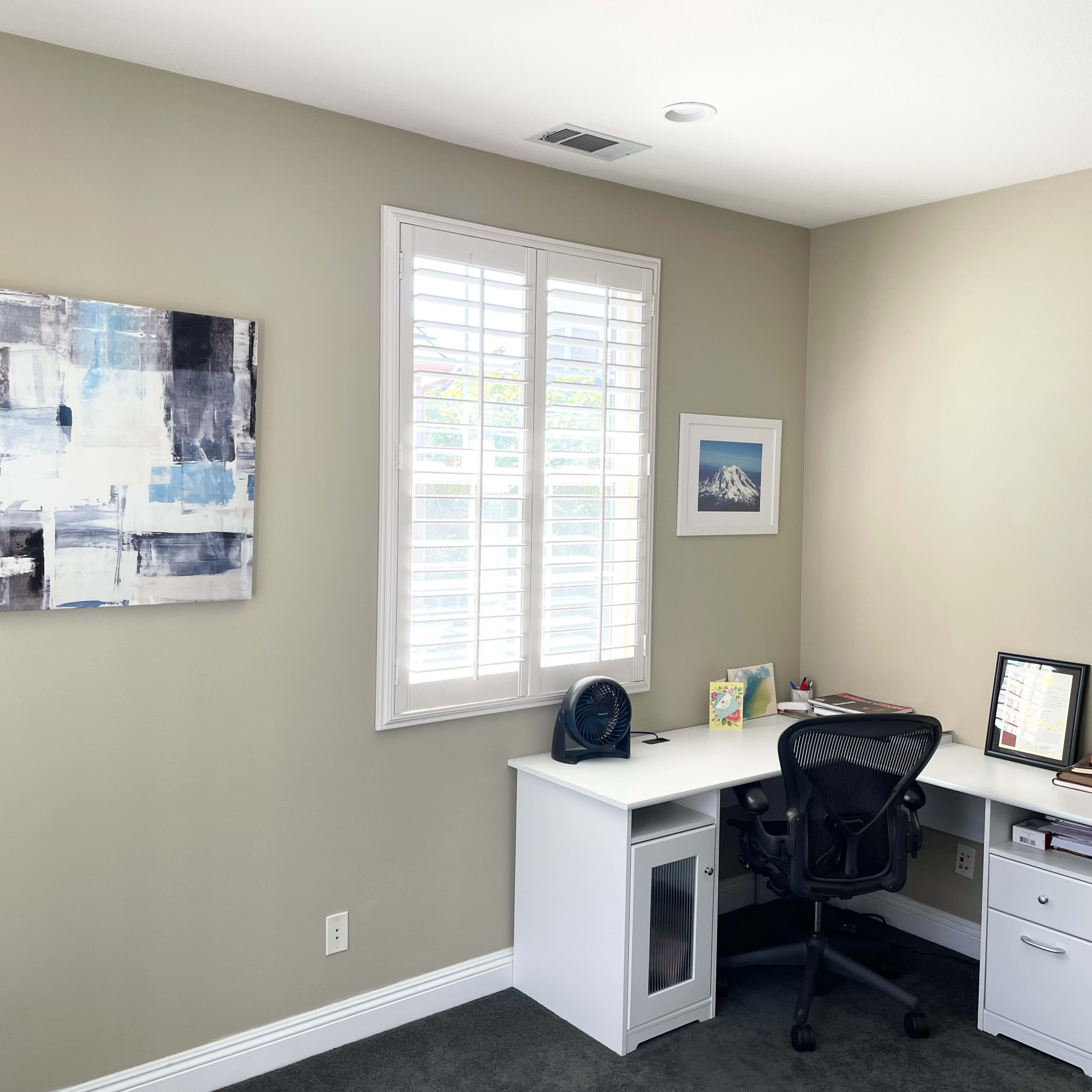 A well-lit office space featuring a white desk with a black swivel chair, a framed photo on the wall, a globe, and an abstract painting hung above the desk.
