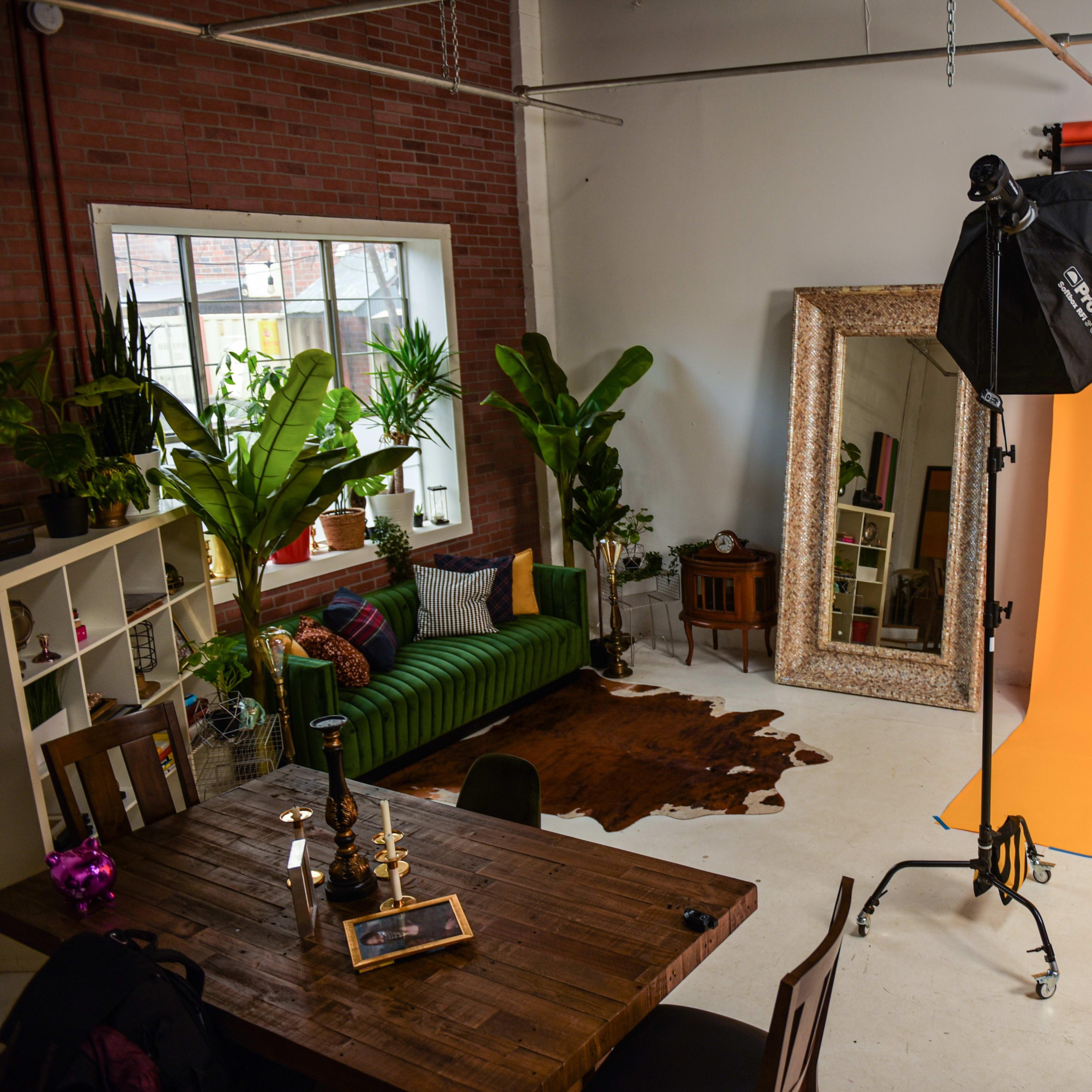 A photography studio featuring an orange backdrop, a green striped couch, a large mirror, tall plants, and a wooden table with decorative items.