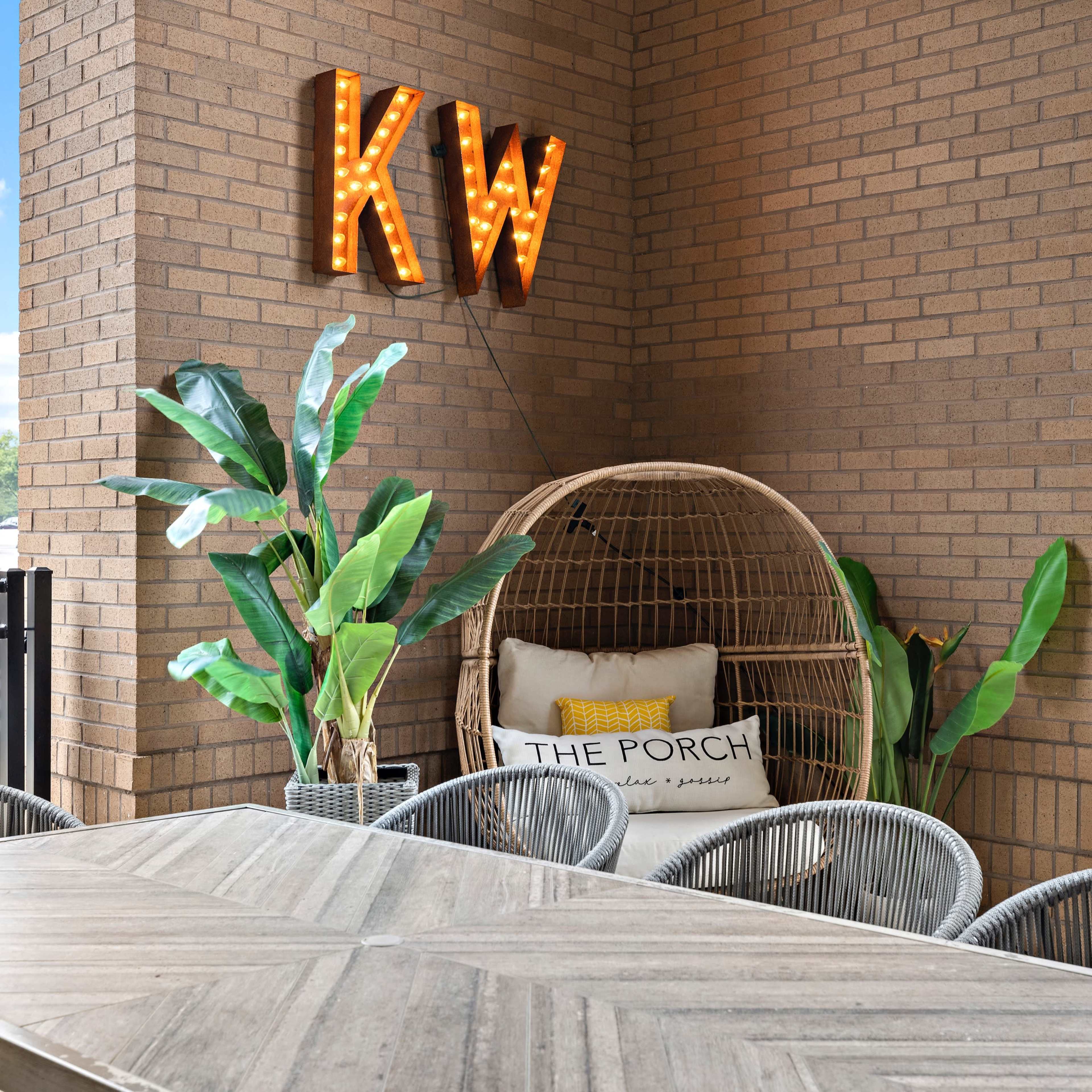 A cozy outdoor seating area features a large table, decorative plants, and a rattan chair with a cushion, alongside a brick wall adorned with illuminated letters.