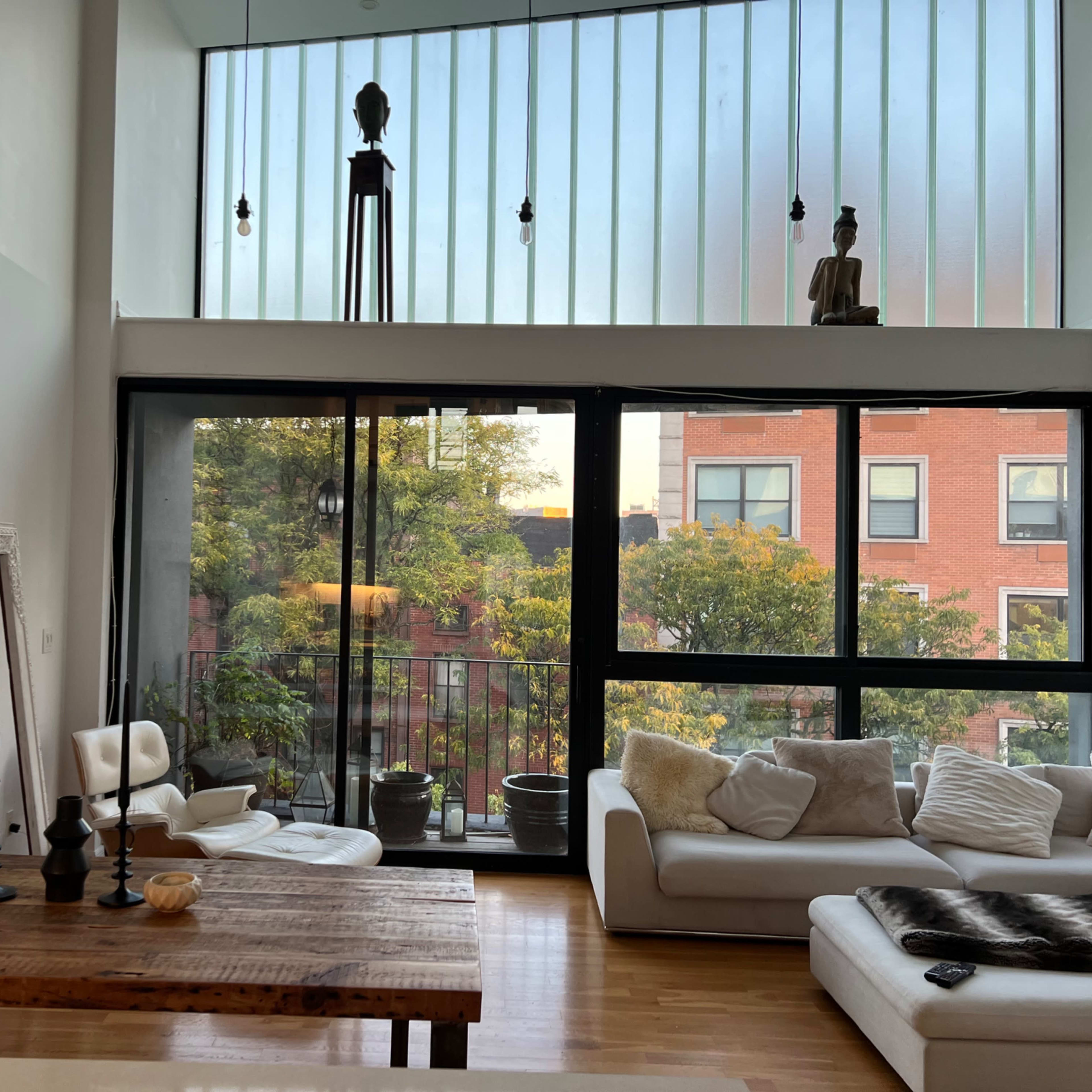 A stylish living room with large windows, a wooden dining table, white furniture, and potted plants.