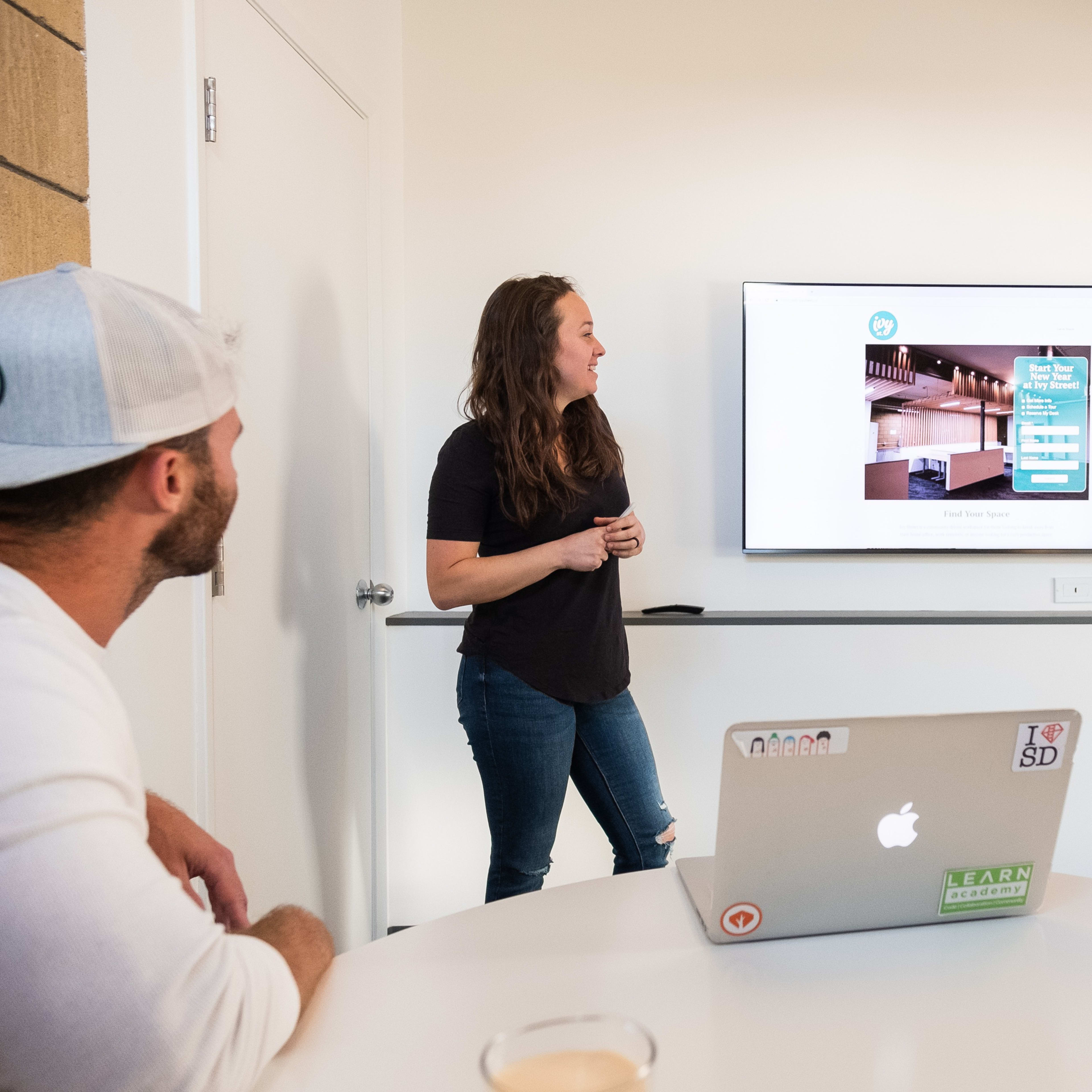 A woman stands by a screen, presenting information to two seated individuals in a meeting room.