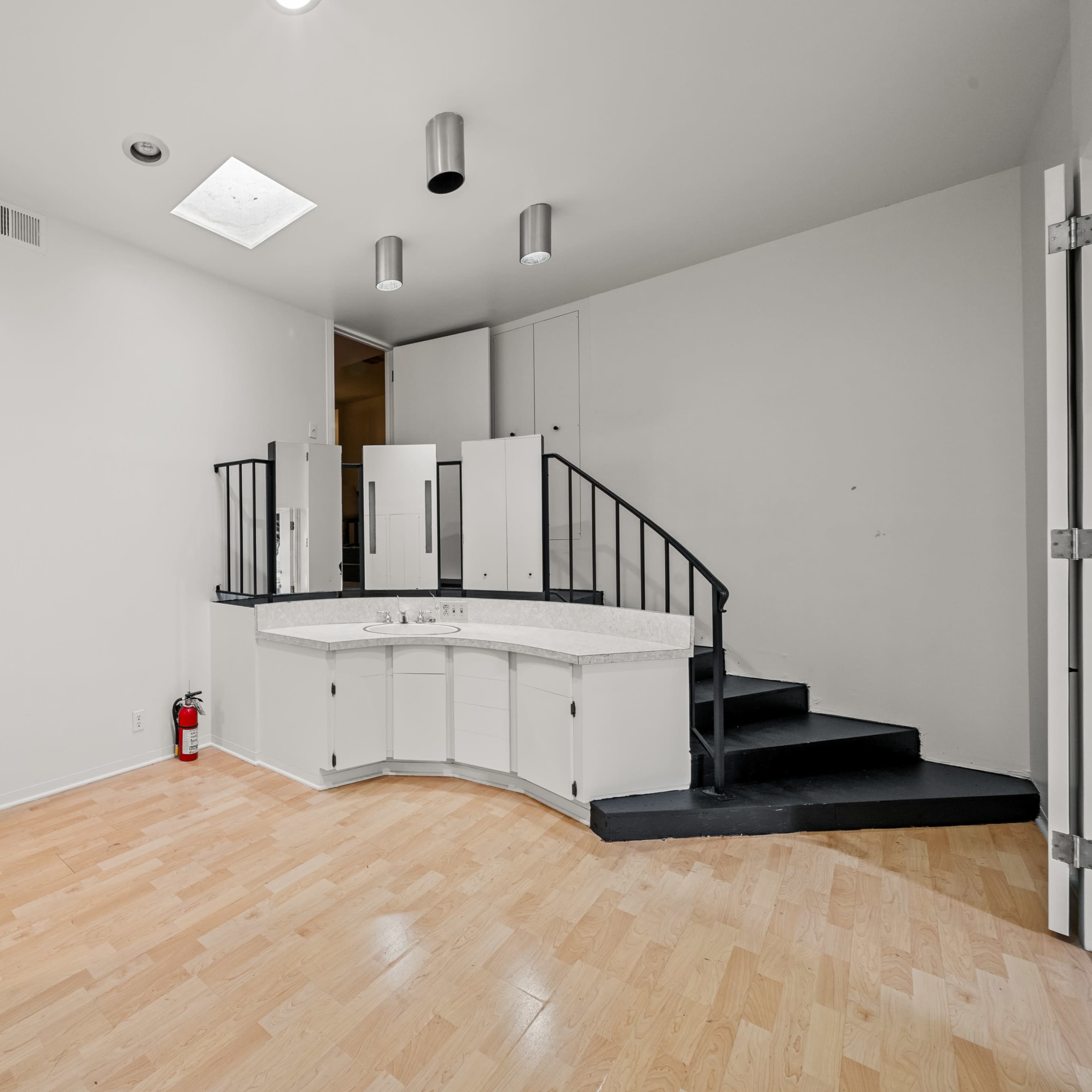 An empty room with light-colored wooden flooring, a curved staircase leading to a higher level, and a small bar area with a countertop.
