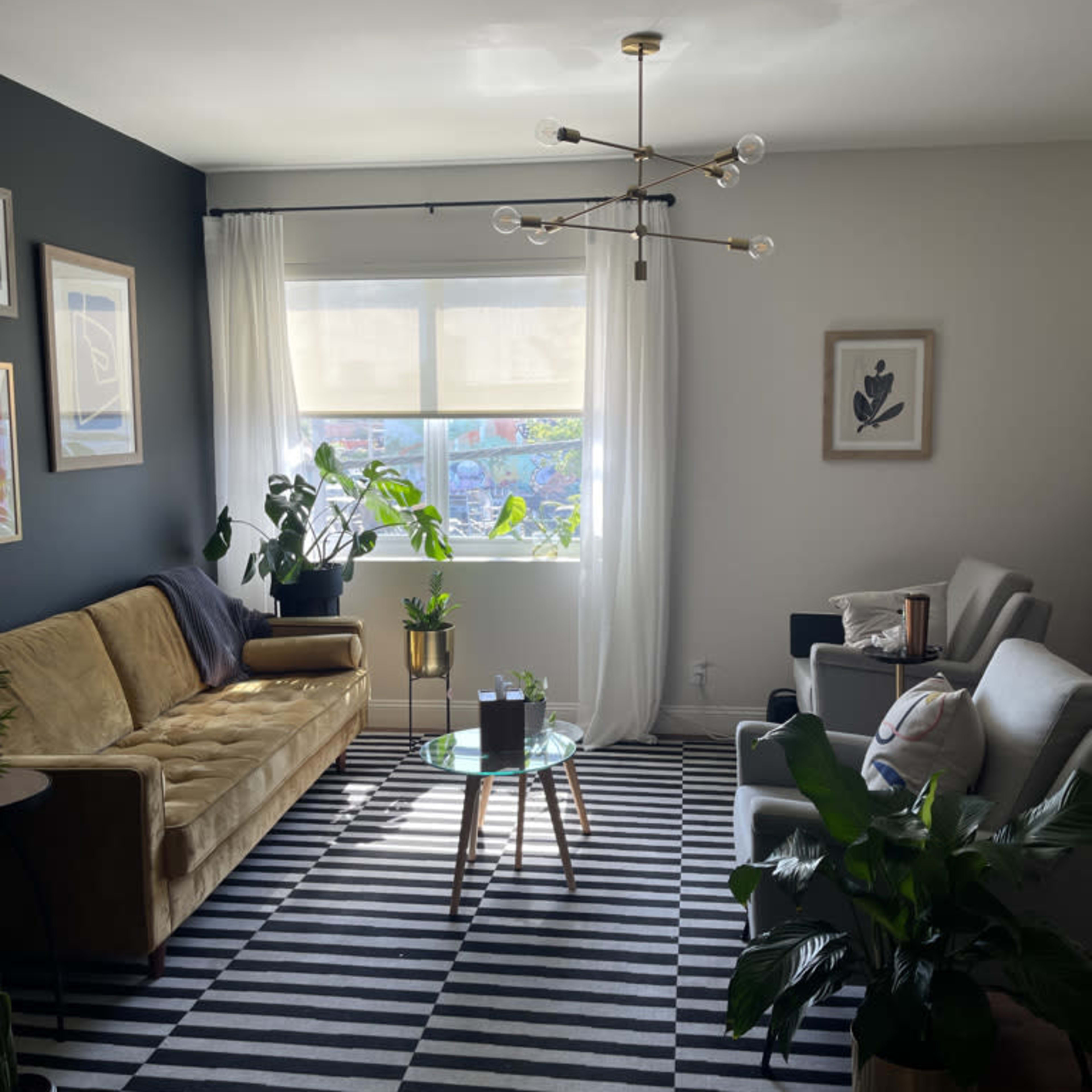 A living room with a mustard-colored sofa, gray armchairs, a striped rug, and various potted plants near a large window.