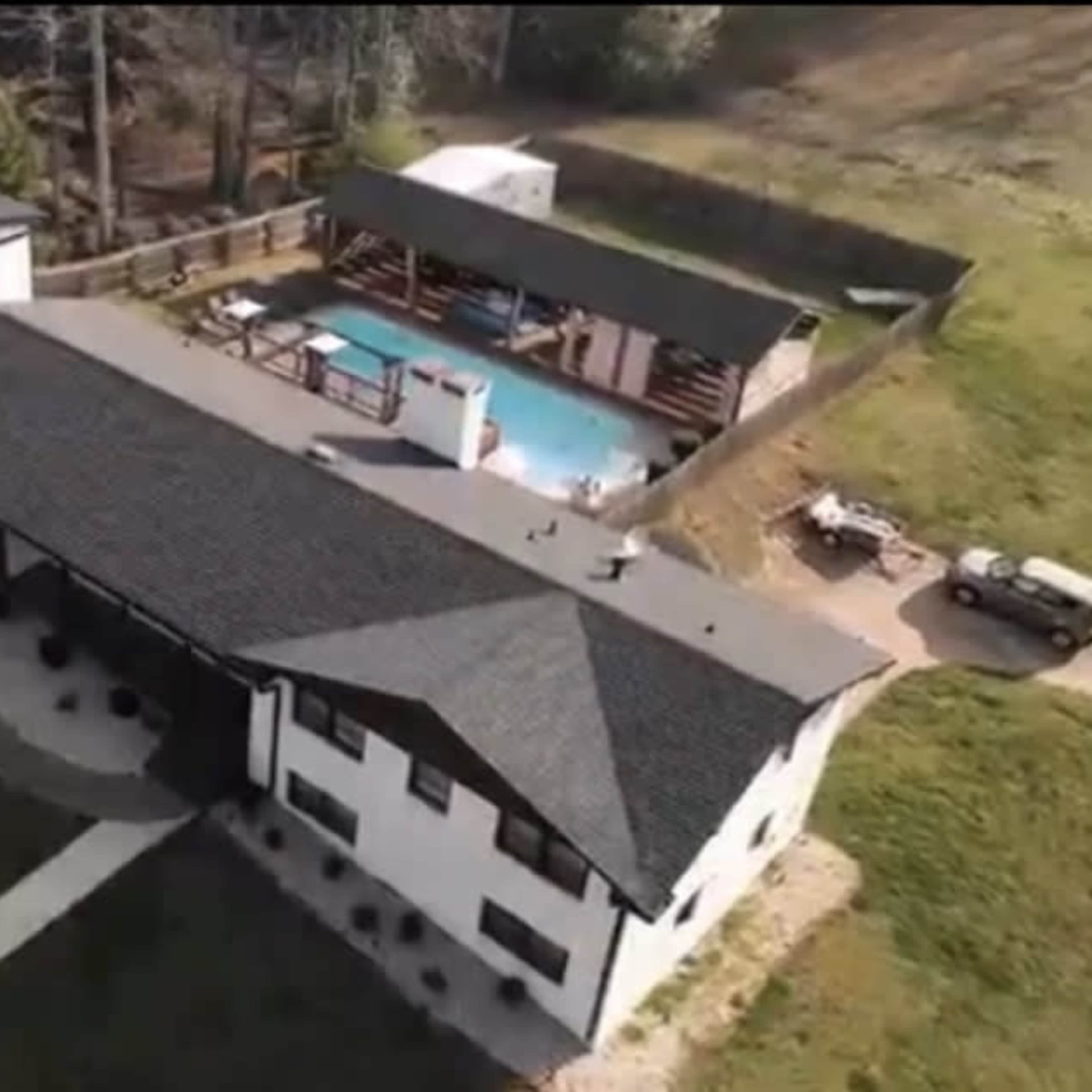 An overhead view of a large modern house with a curved driveway, a swimming pool in the backyard, and several outbuildings.