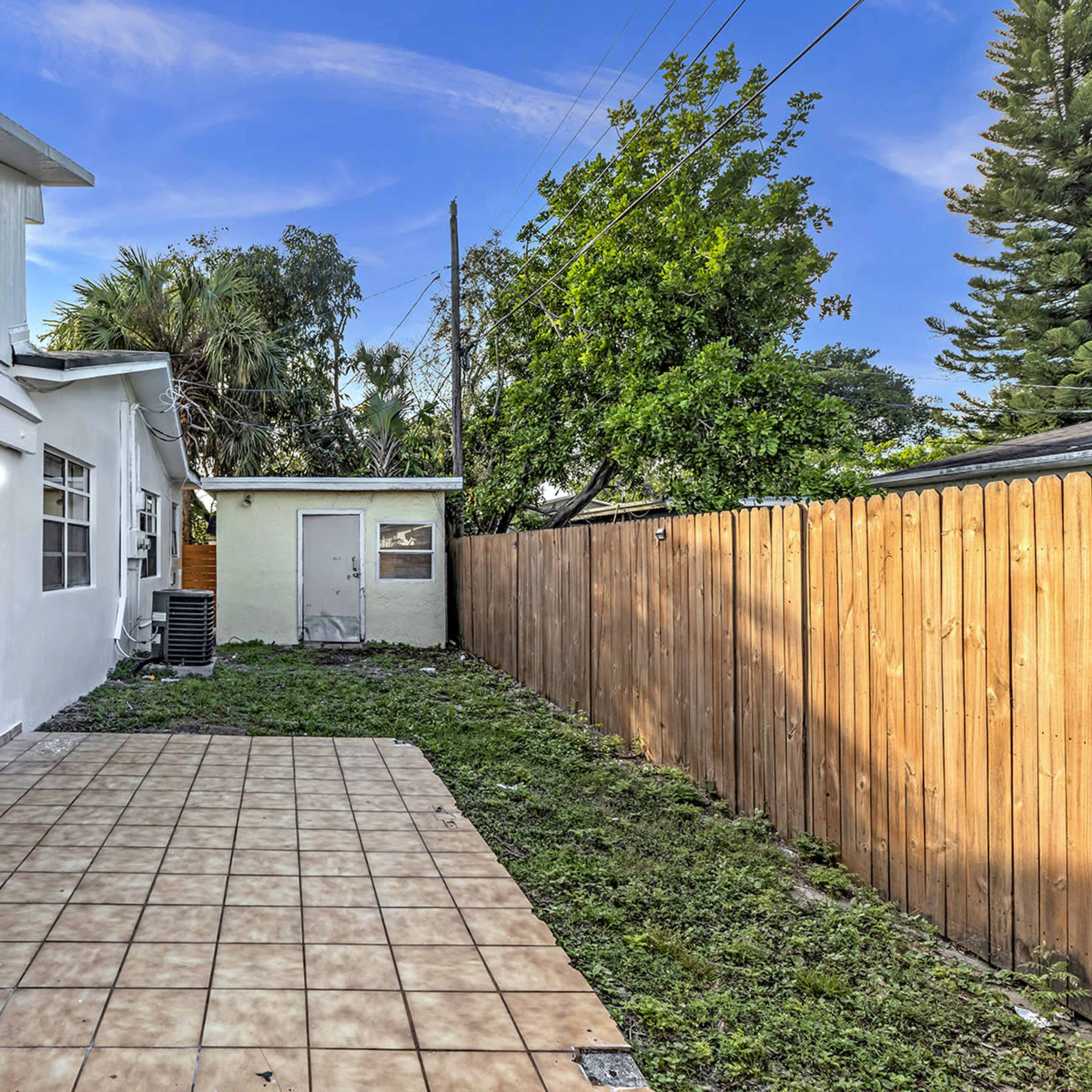 A backyard area with a tiled patio, a small shed, and overgrown grass, bordered by a wooden fence.