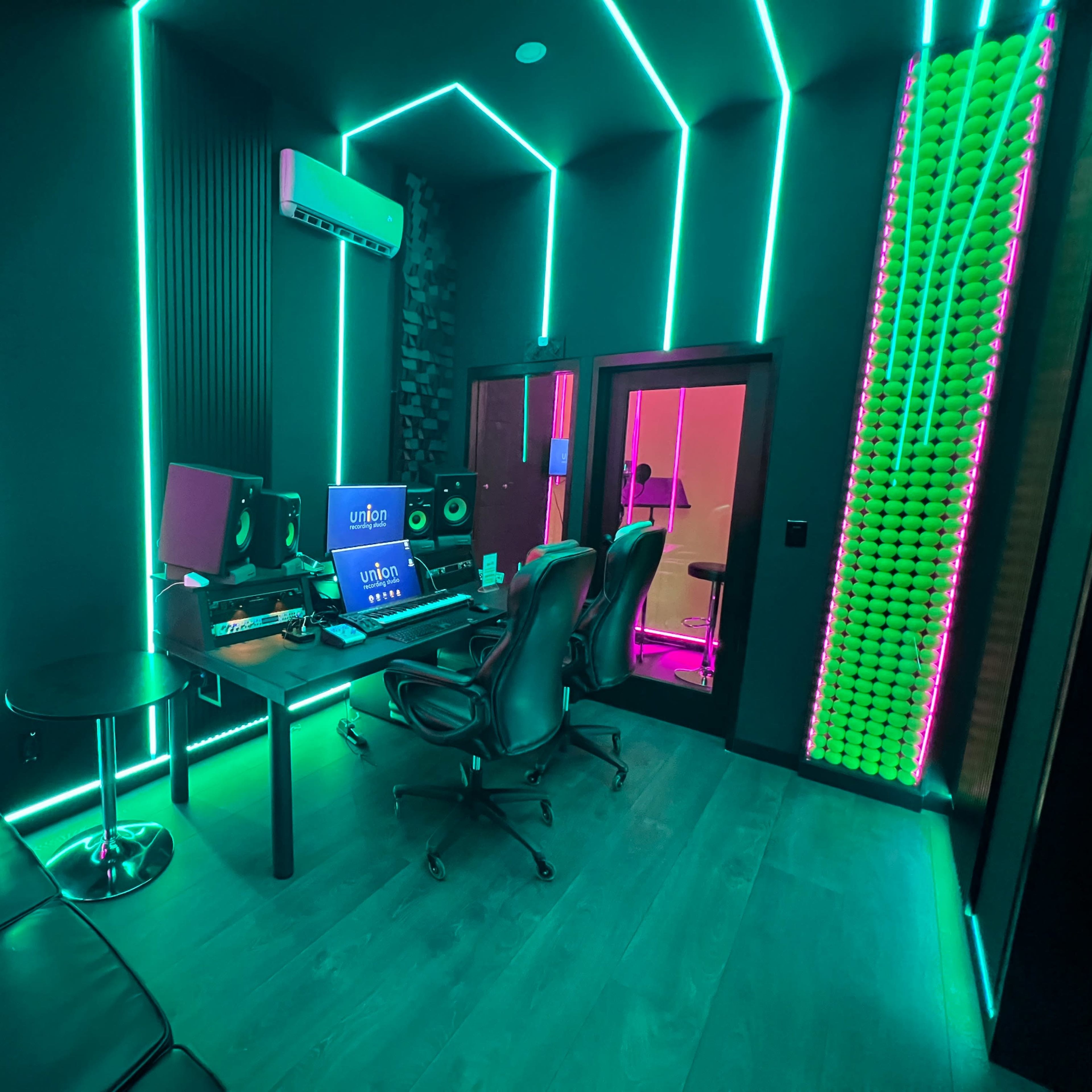 A modern music studio with neon lighting, featuring a desk with computer equipment, two black ergonomic chairs, and a lounge area.