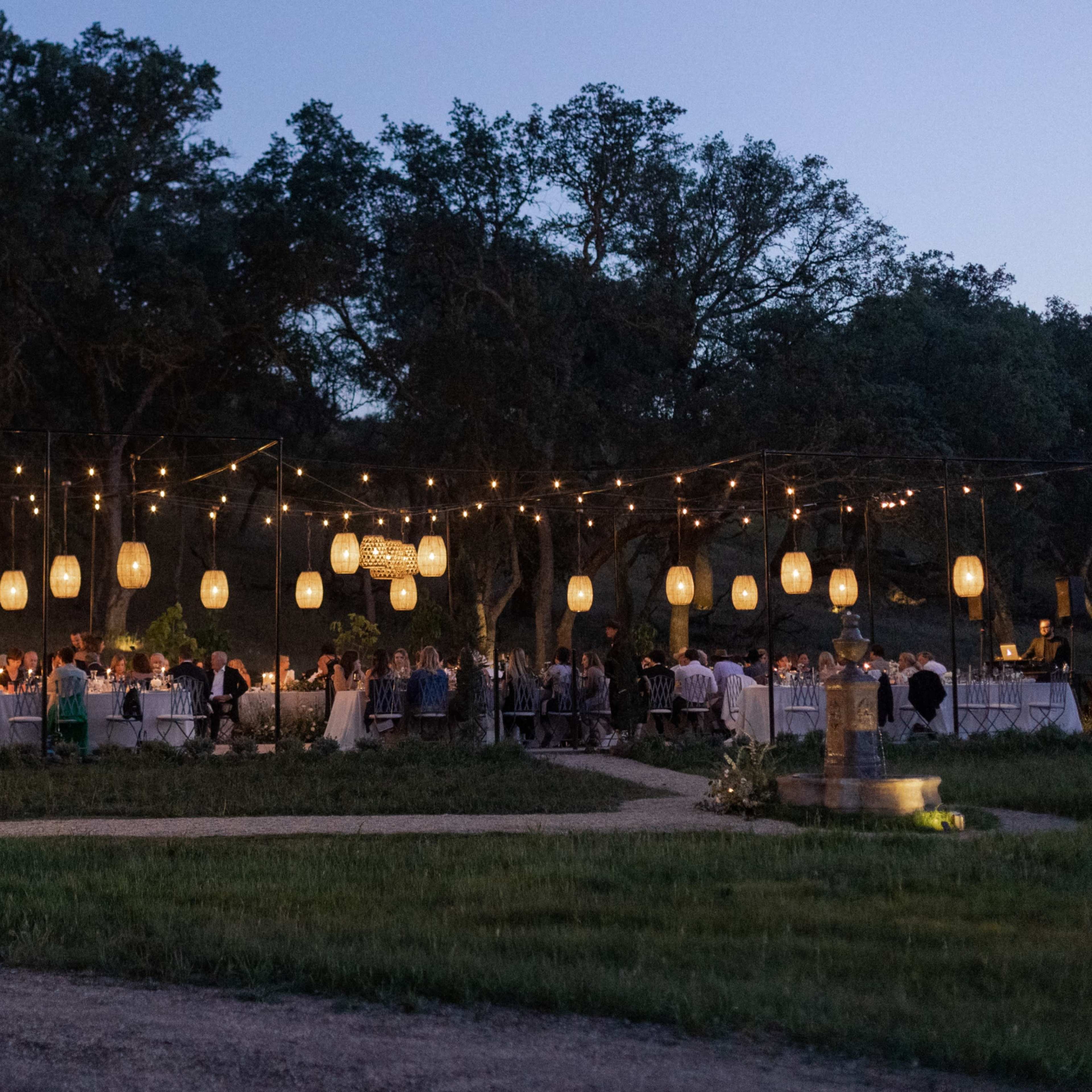 A large outdoor gathering is held at dusk, featuring tables surrounded by guests and illuminated lanterns hanging above.