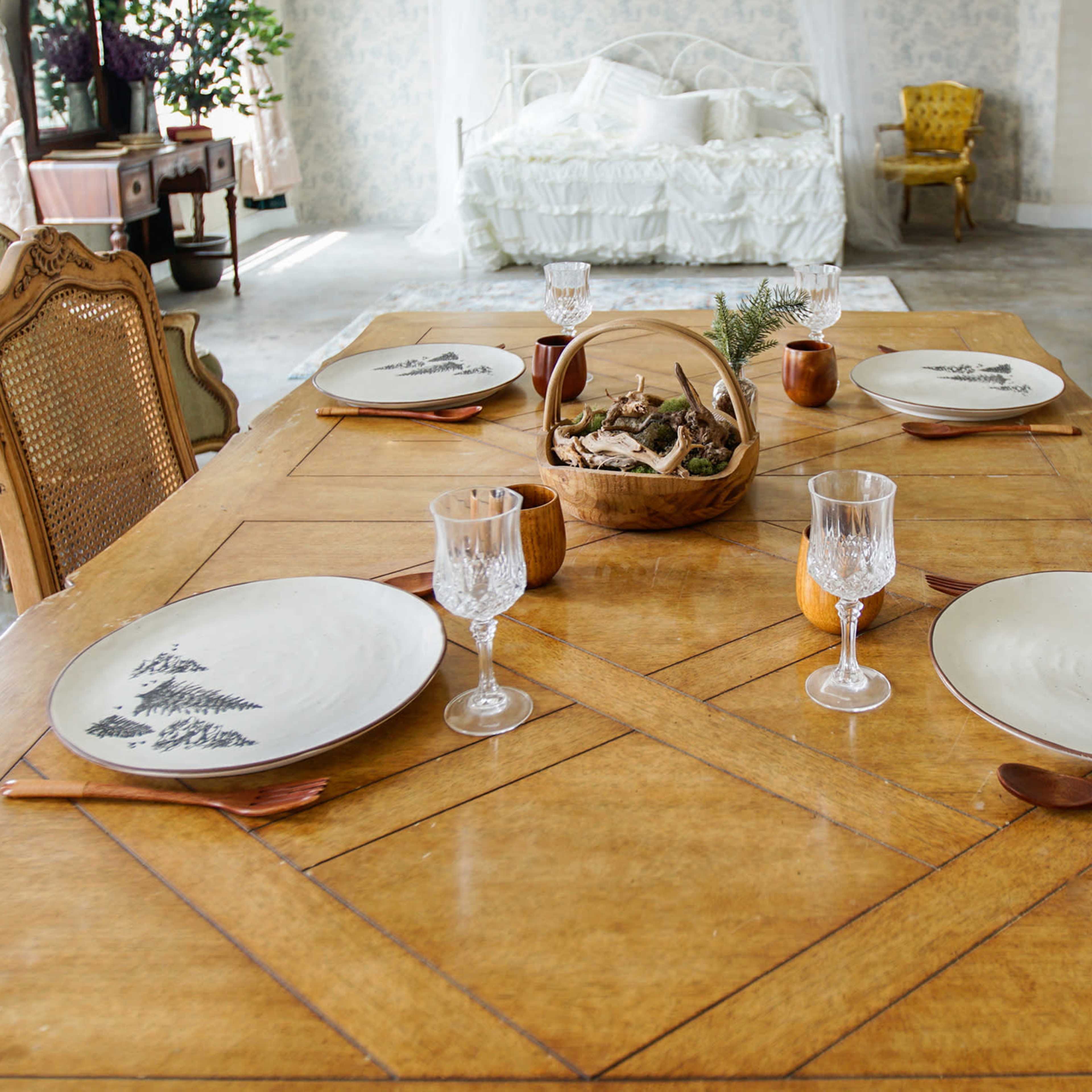 A wooden dining table is set with four plates, crystal glasses, and a decorative bowl filled with natural materials, surrounded by a light-filled room with vintage furniture.