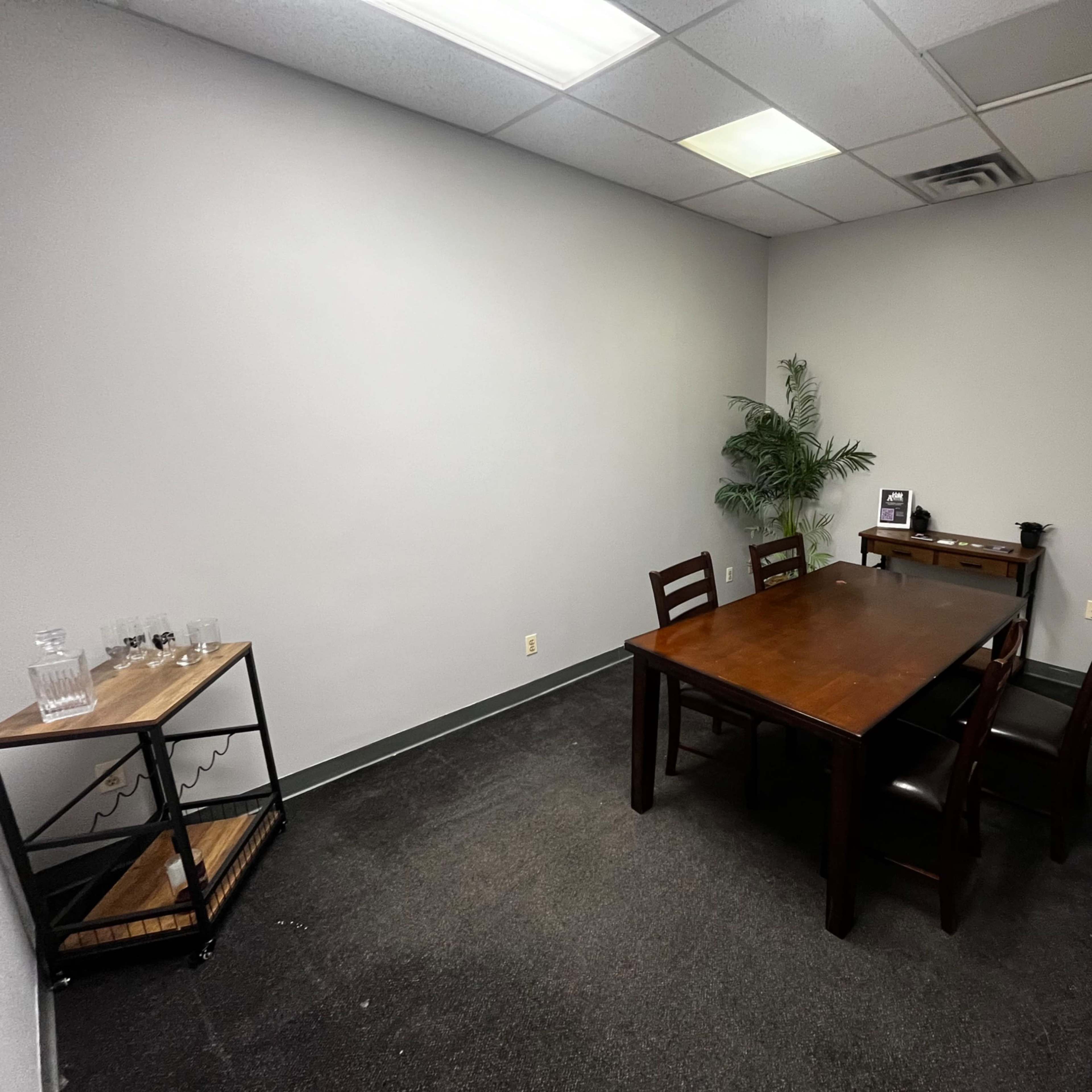A sparsely furnished meeting room with a wooden table, four chairs, a small shelving unit, and a potted plant in the corner.