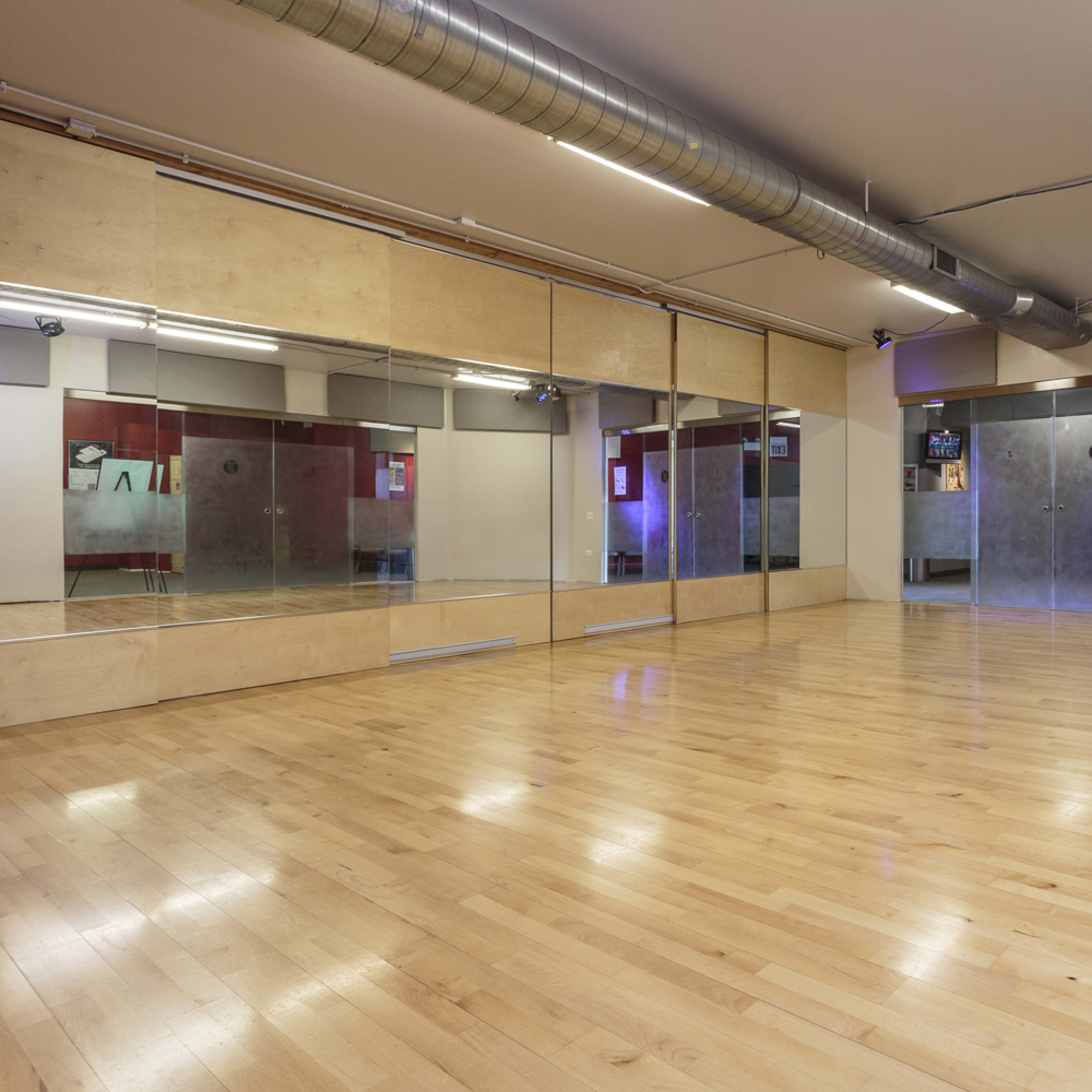 A dance studio features wooden flooring and large mirrors on one wall, with a minimalist design and dim lighting.
