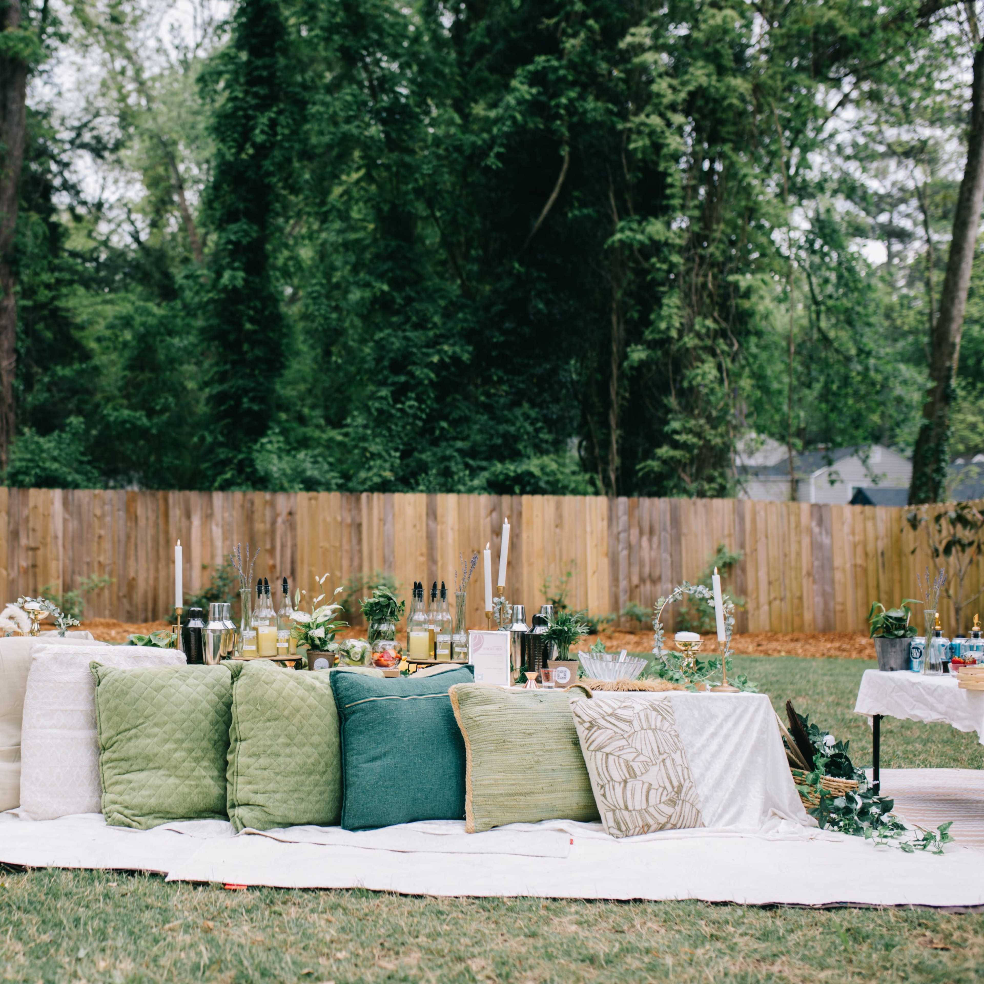 A picnic setup with green and white pillows, a white blanket, and various food and drink items is displayed on a grassy area surrounded by a wooden fence and trees.