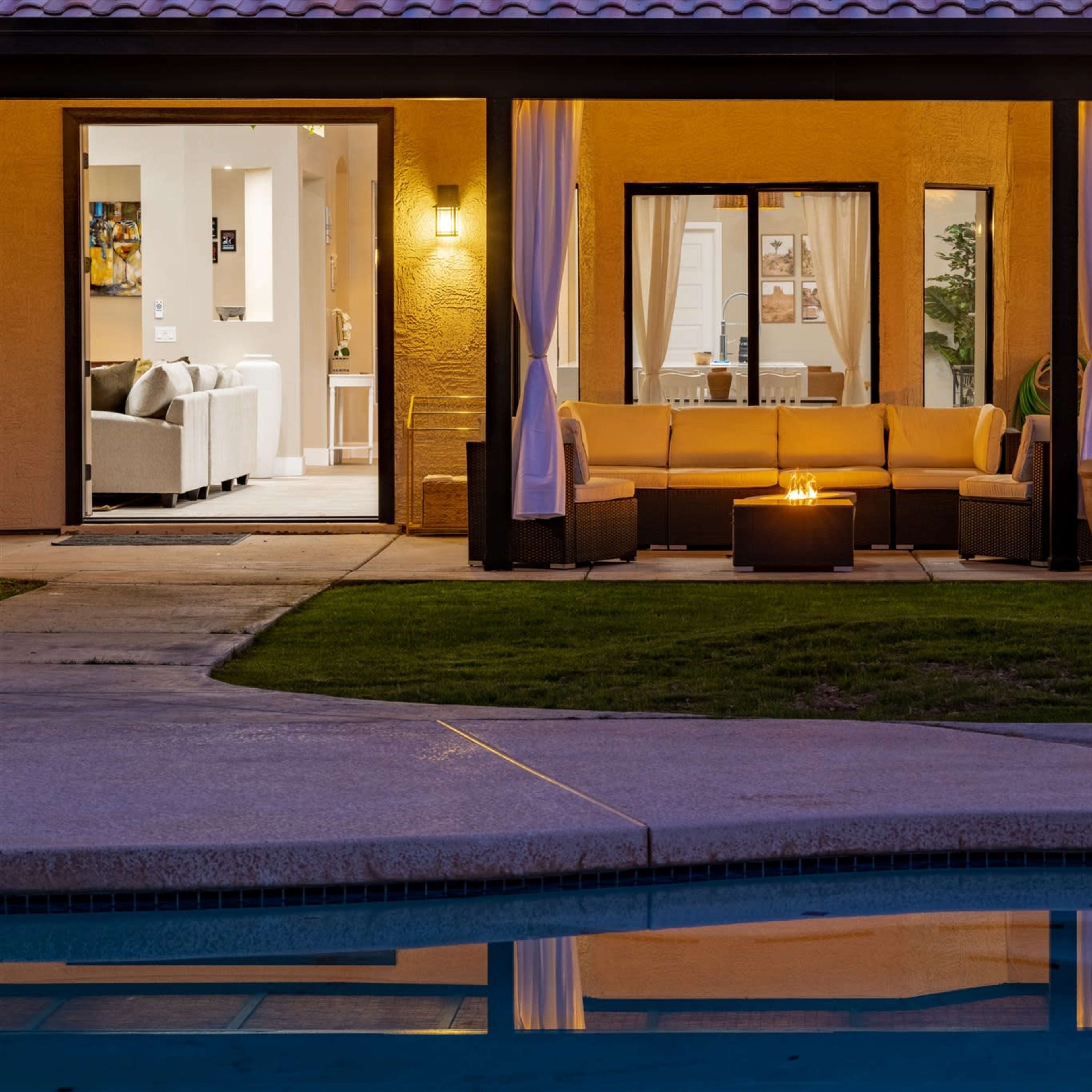 A modern outdoor lounge area with a fire pit is situated beside a swimming pool, reflecting the lights of the adjacent indoor living space.