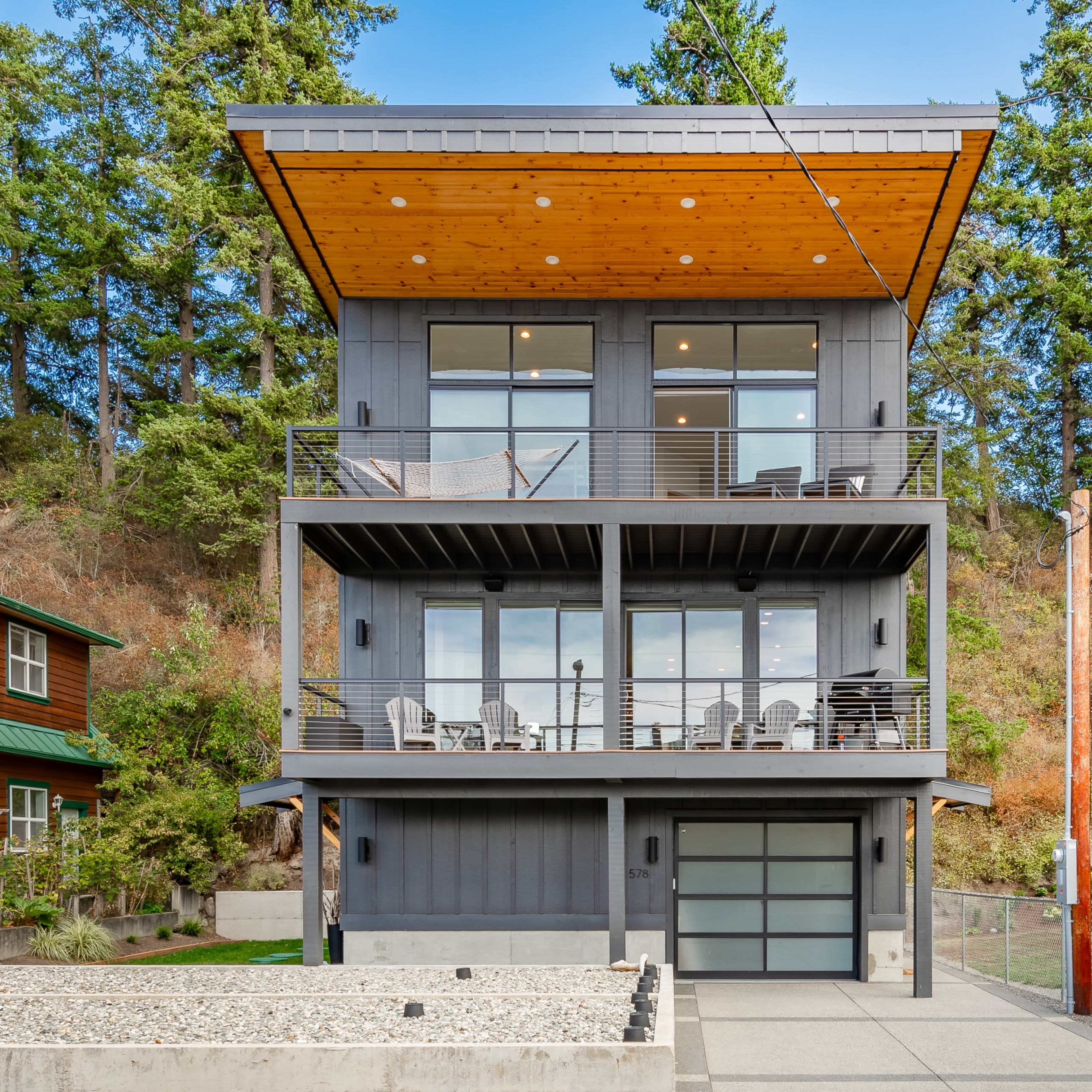 A modern three-story house with large windows, a wooden overhang, and a concrete driveway, surrounded by trees.