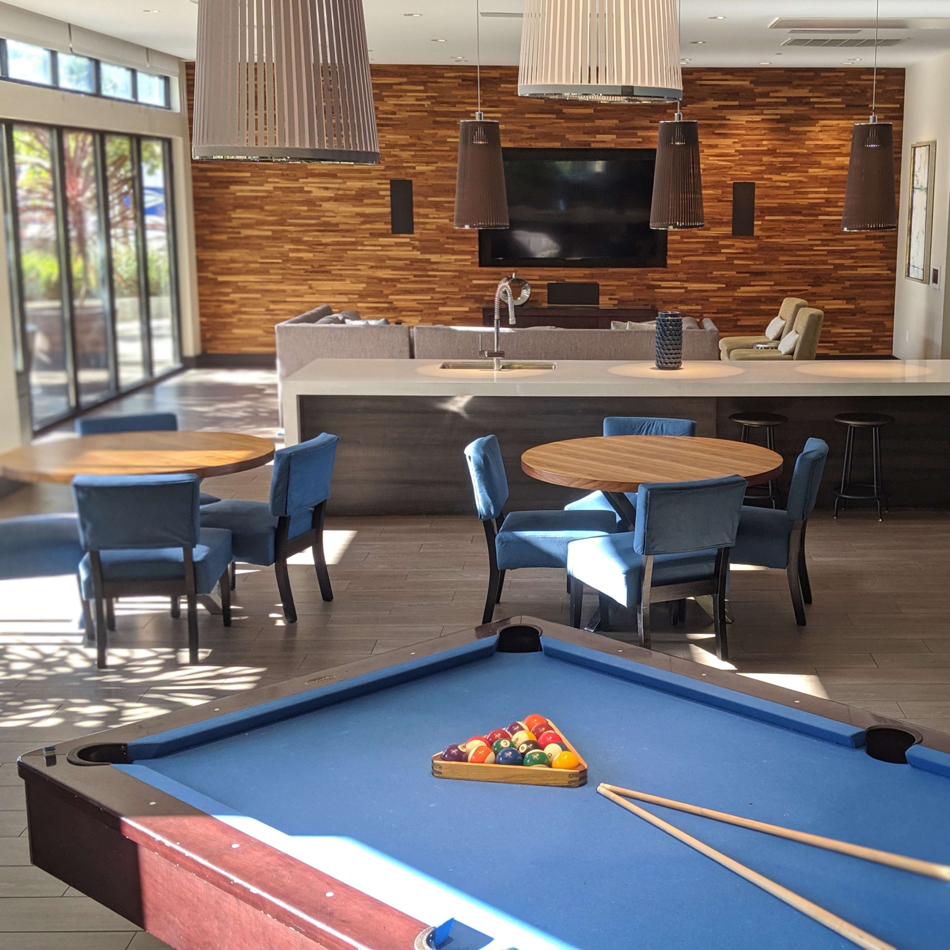 A modern lounge area featuring a billiard table in the foreground, round tables with blue chairs, and a stone wall with a television in the background.