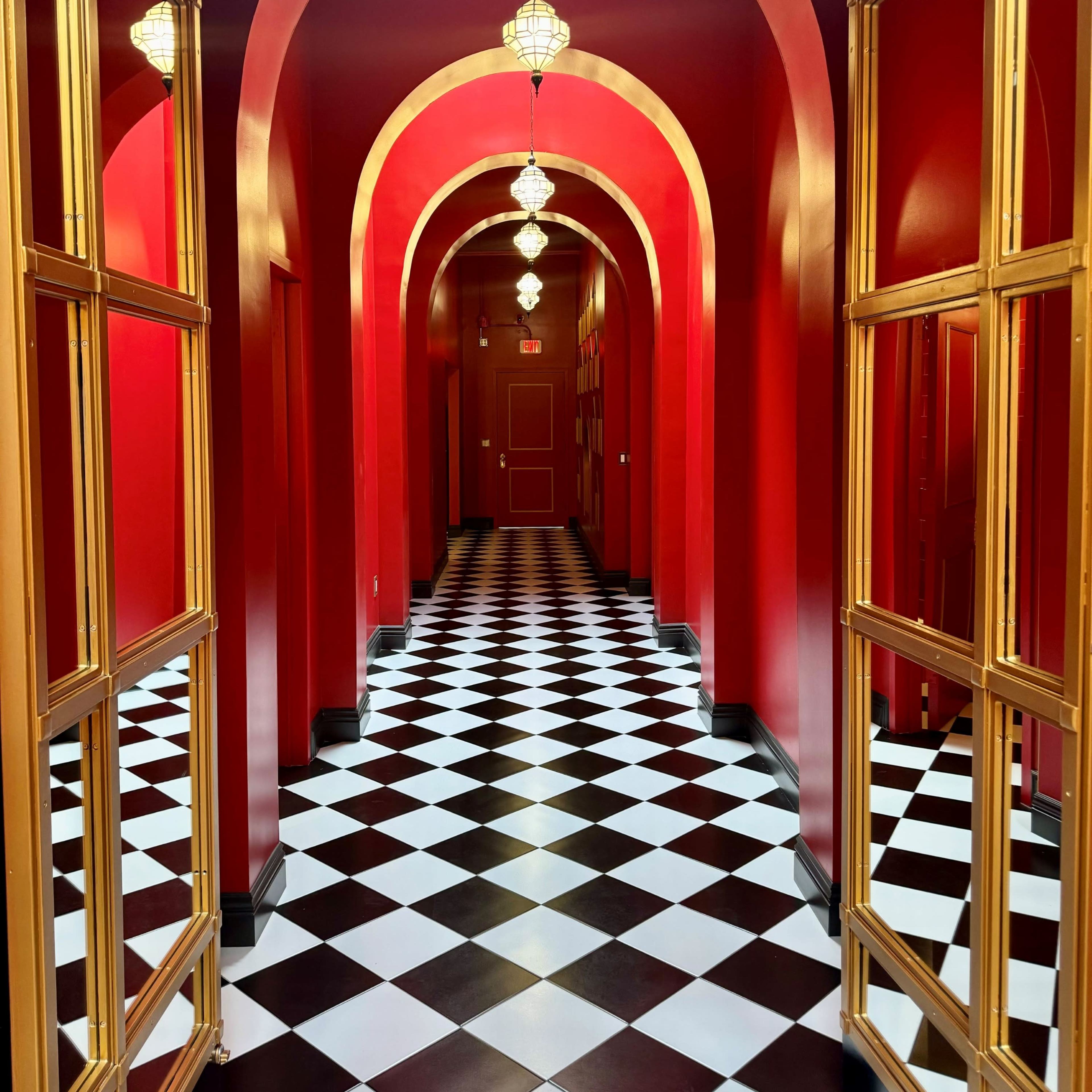 A corridor with red walls and arches, featuring a black and white checkered floor and golden-framed mirrors leading into the distance.