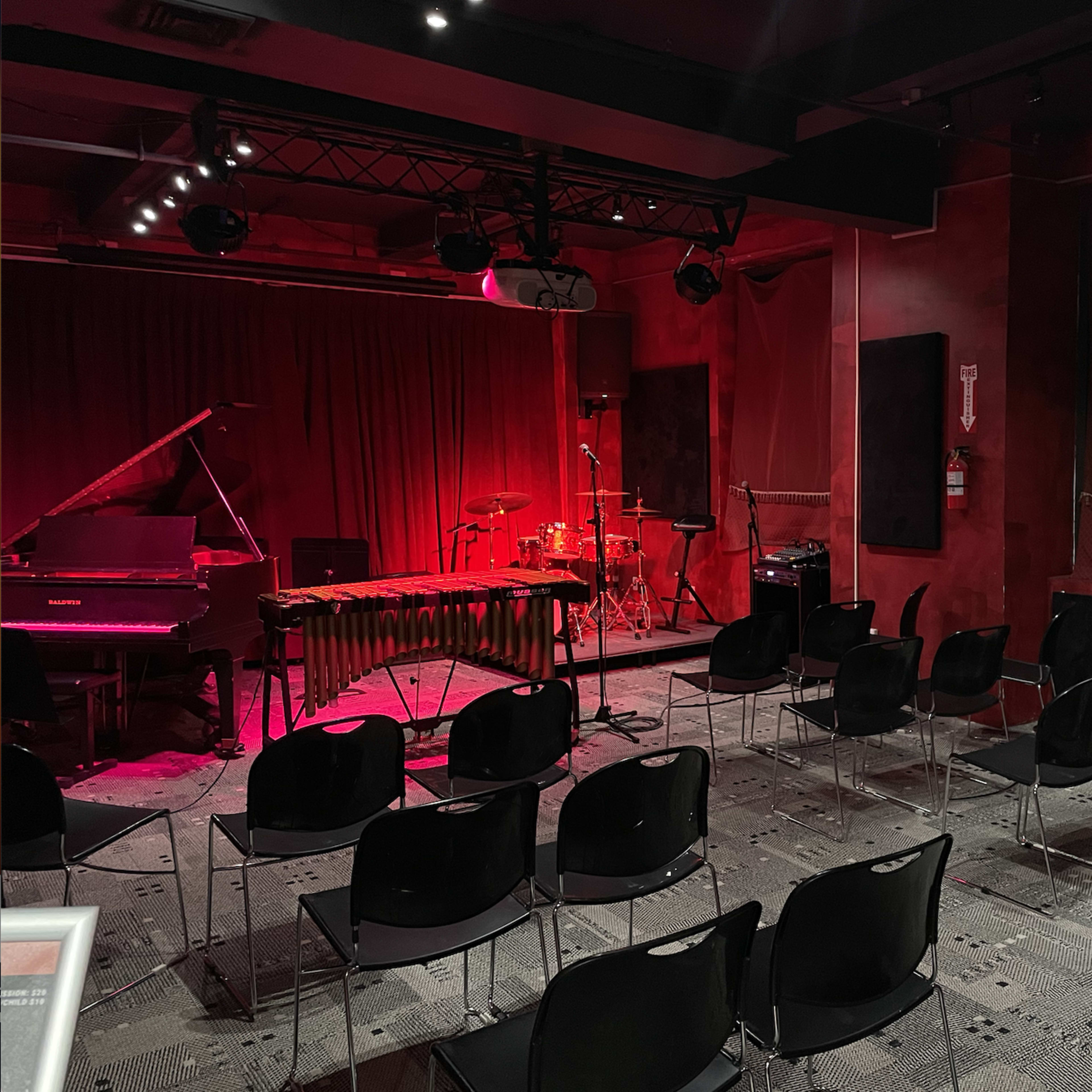 A small music performance venue features a stage with a piano, vibraphone, and drum set, surrounded by rows of empty black chairs.