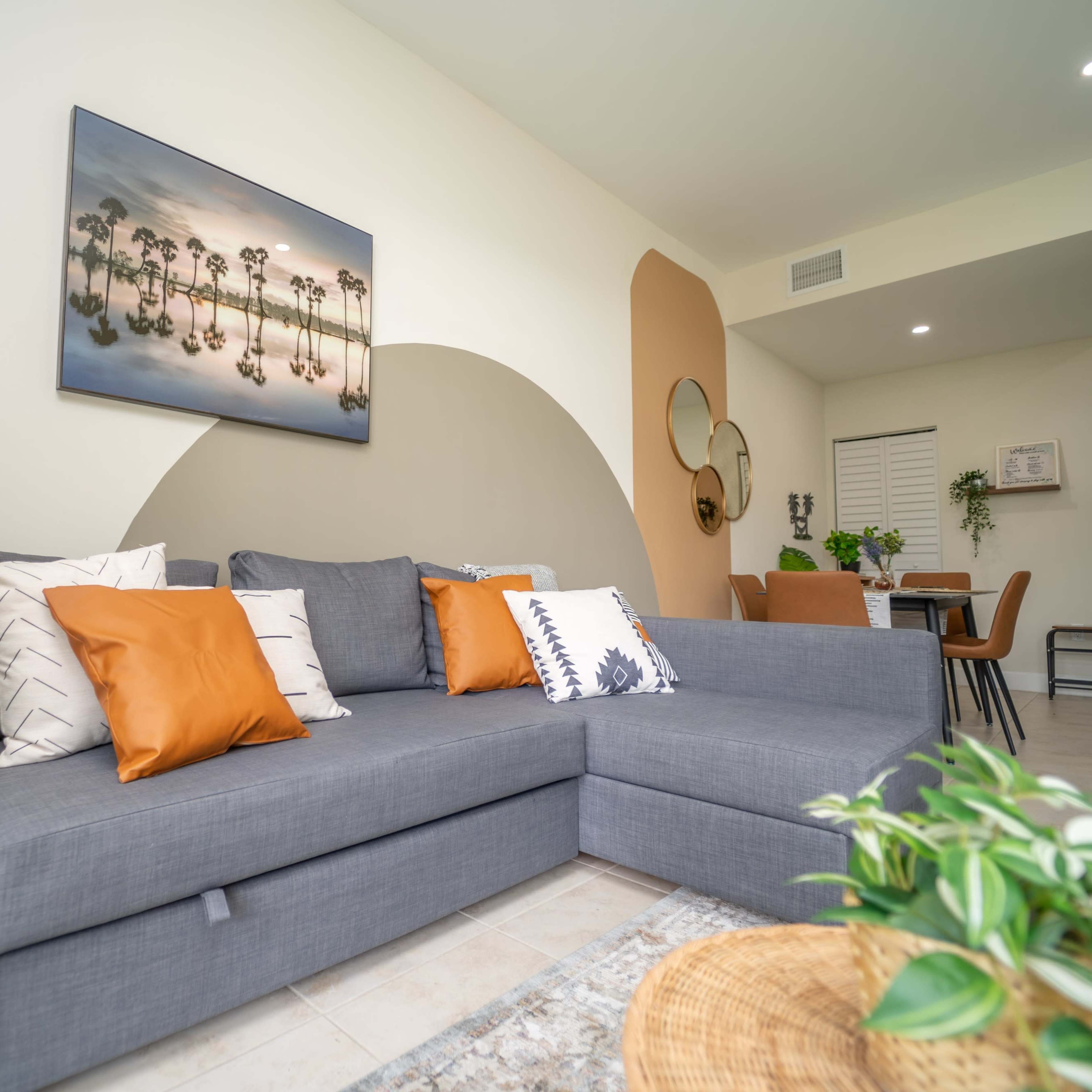 A modern living room with a gray sectional sofa adorned with orange and patterned pillows, a large outdoor-themed artwork on the wall, and an open layout leading to a dining area.