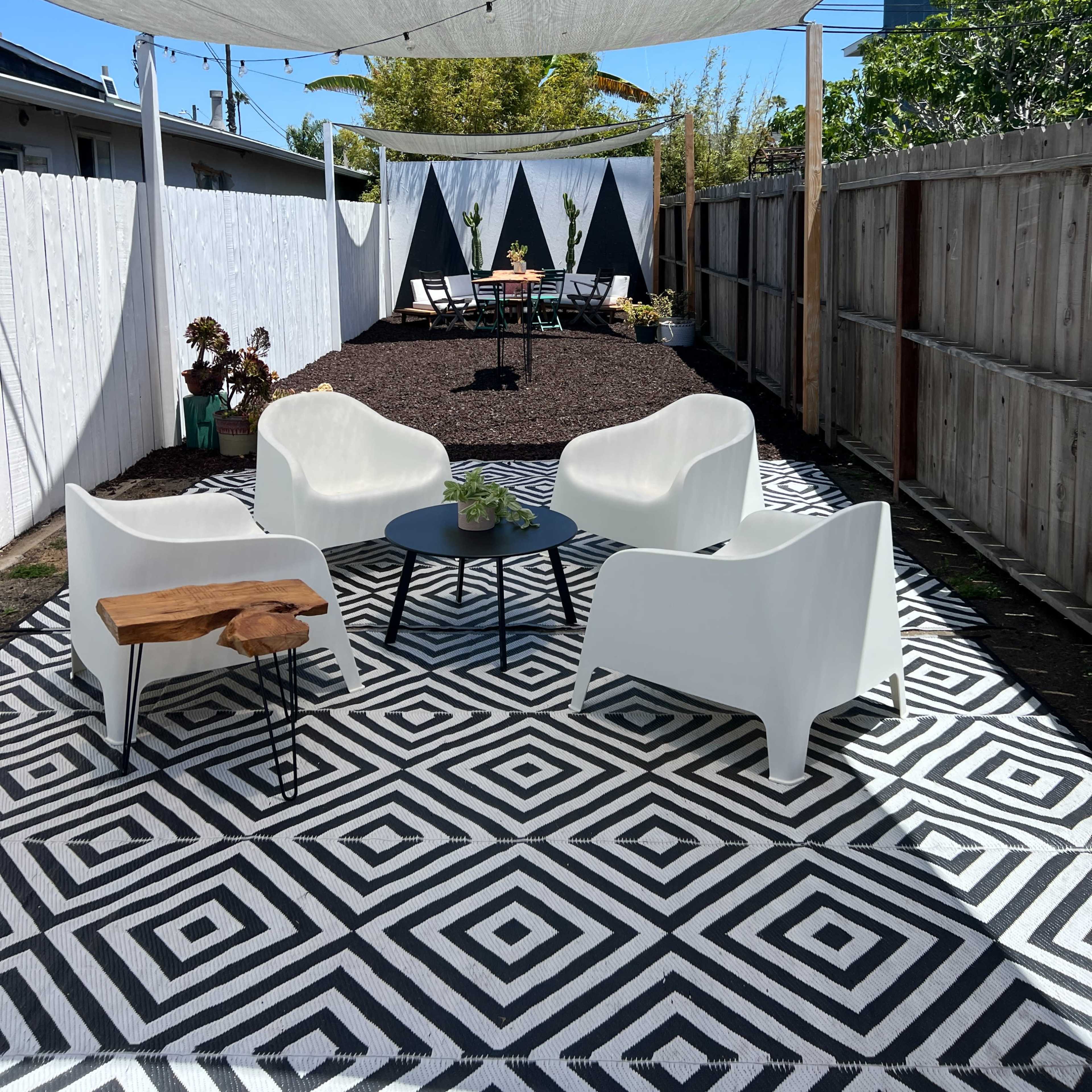 A patio area features white chairs arranged around a circular table on a geometric-patterned rug, with a shaded section and a backdrop of decorative fabric triangles.