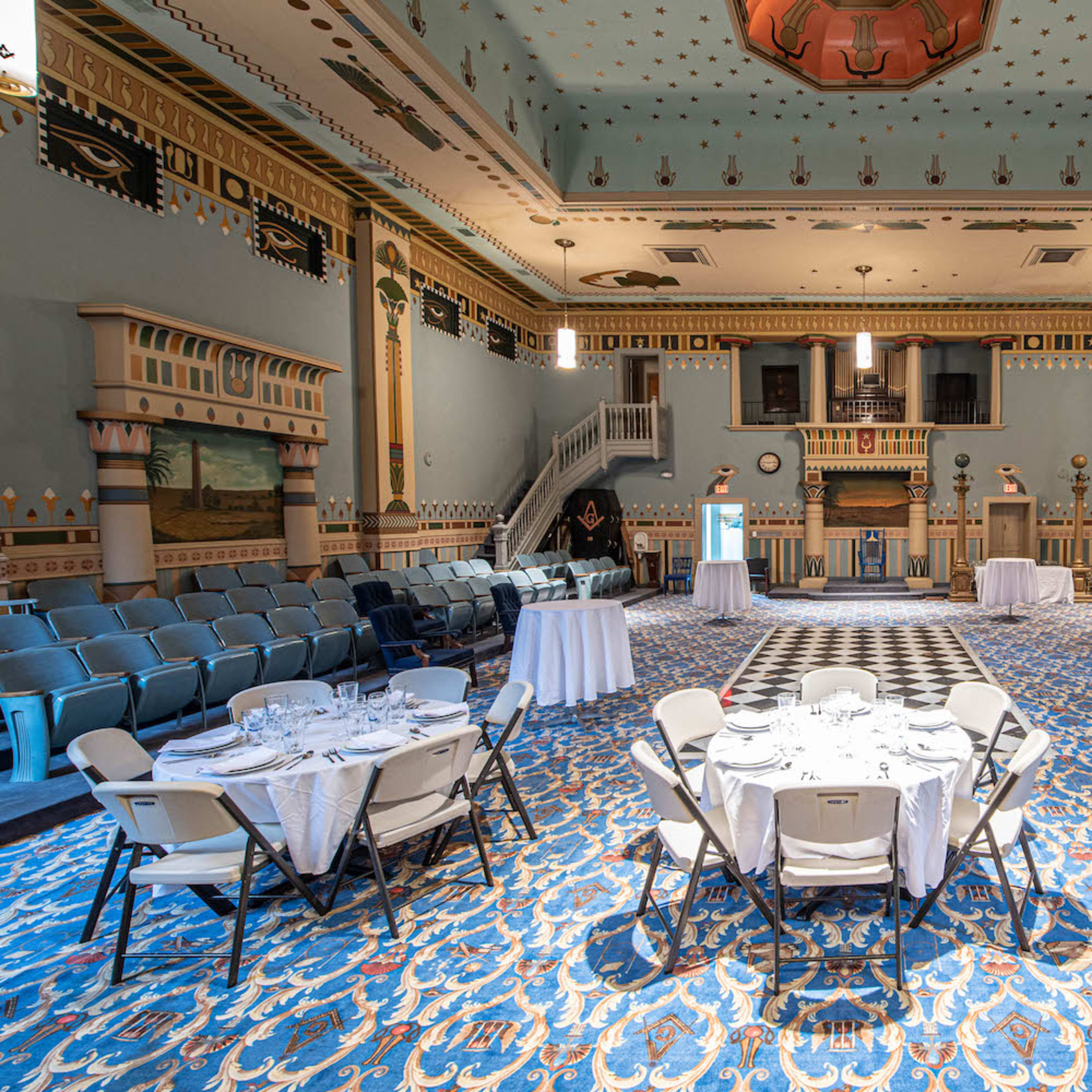 A spacious, elegantly decorated hall with circular tables set for dining, surrounded by rows of chairs and ornate walls.