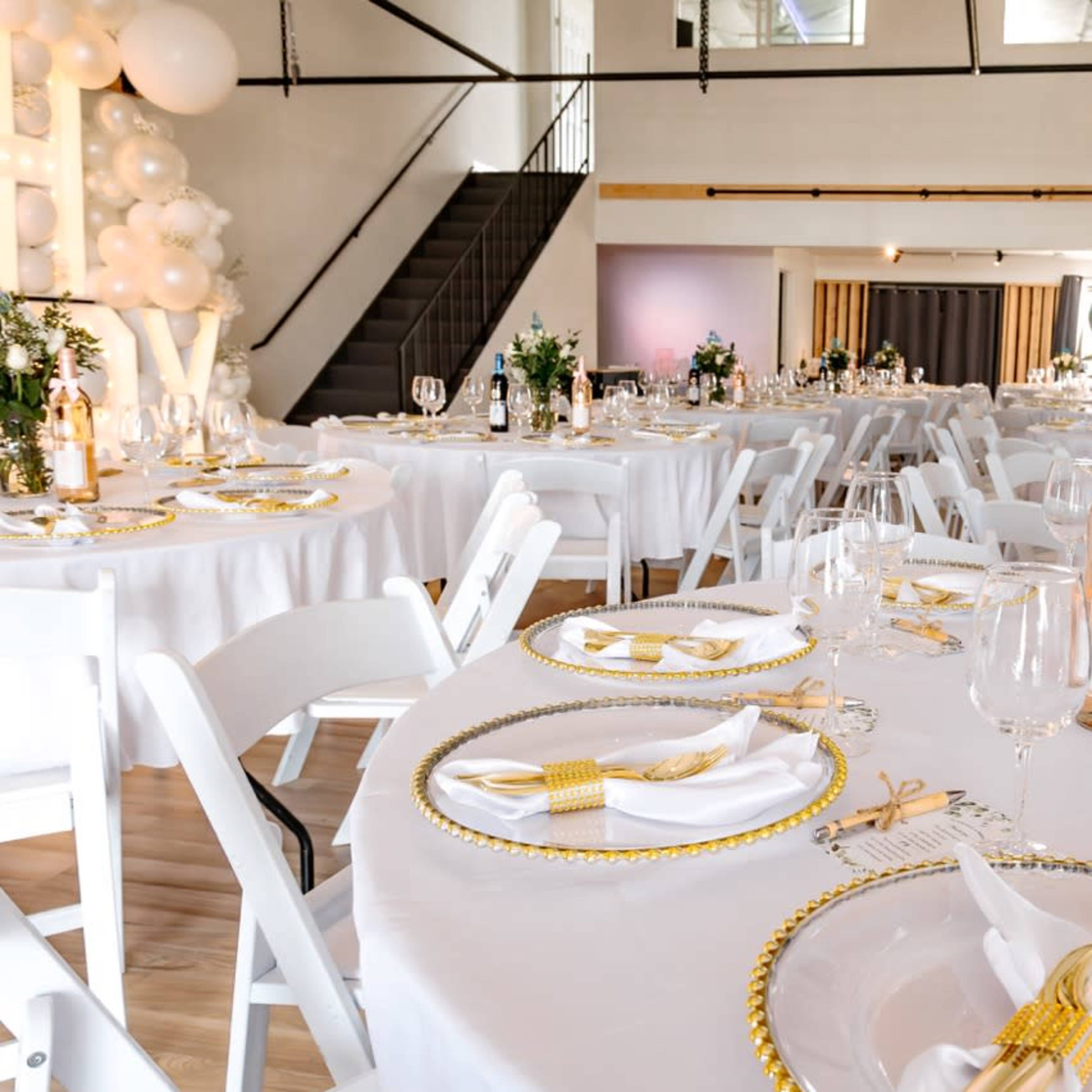 A beautifully arranged event space with round tables set with white tablecloths, golden accents, and floral centerpieces.