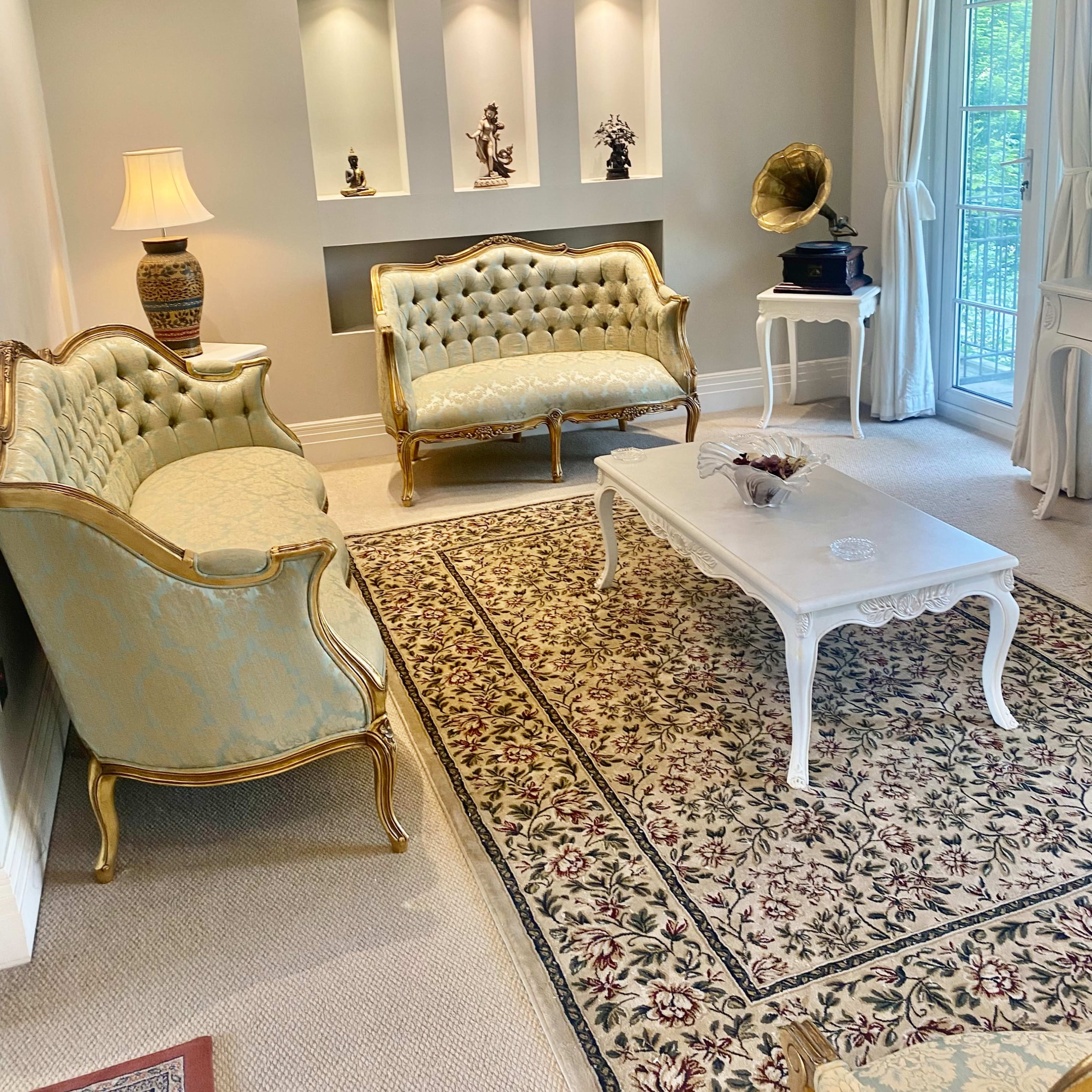 A light-colored living room with two ornate sofas, a decorative coffee table, and a patterned area rug.