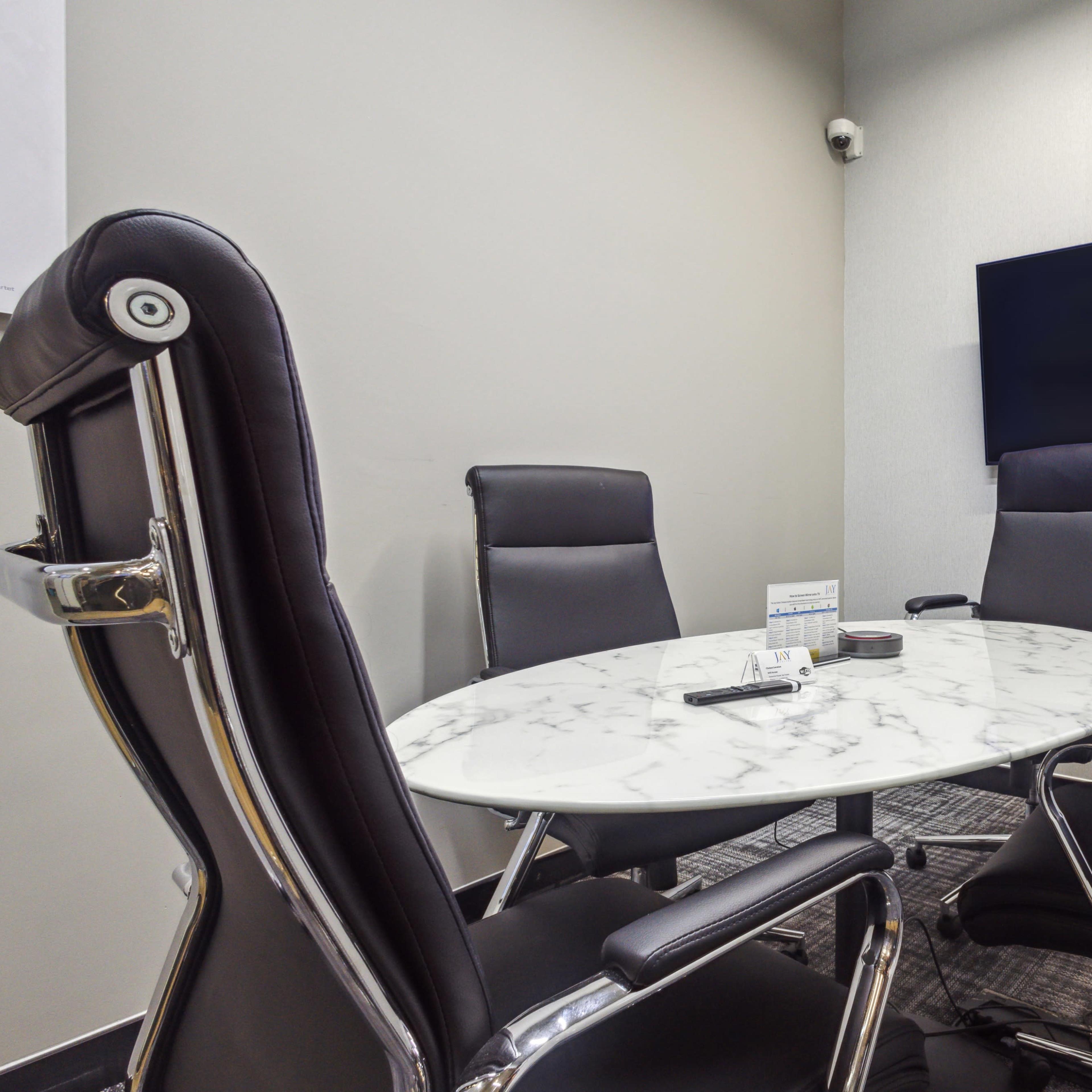 A small, modern conference room features a round marble table surrounded by four black executive chairs and a wall-mounted TV.