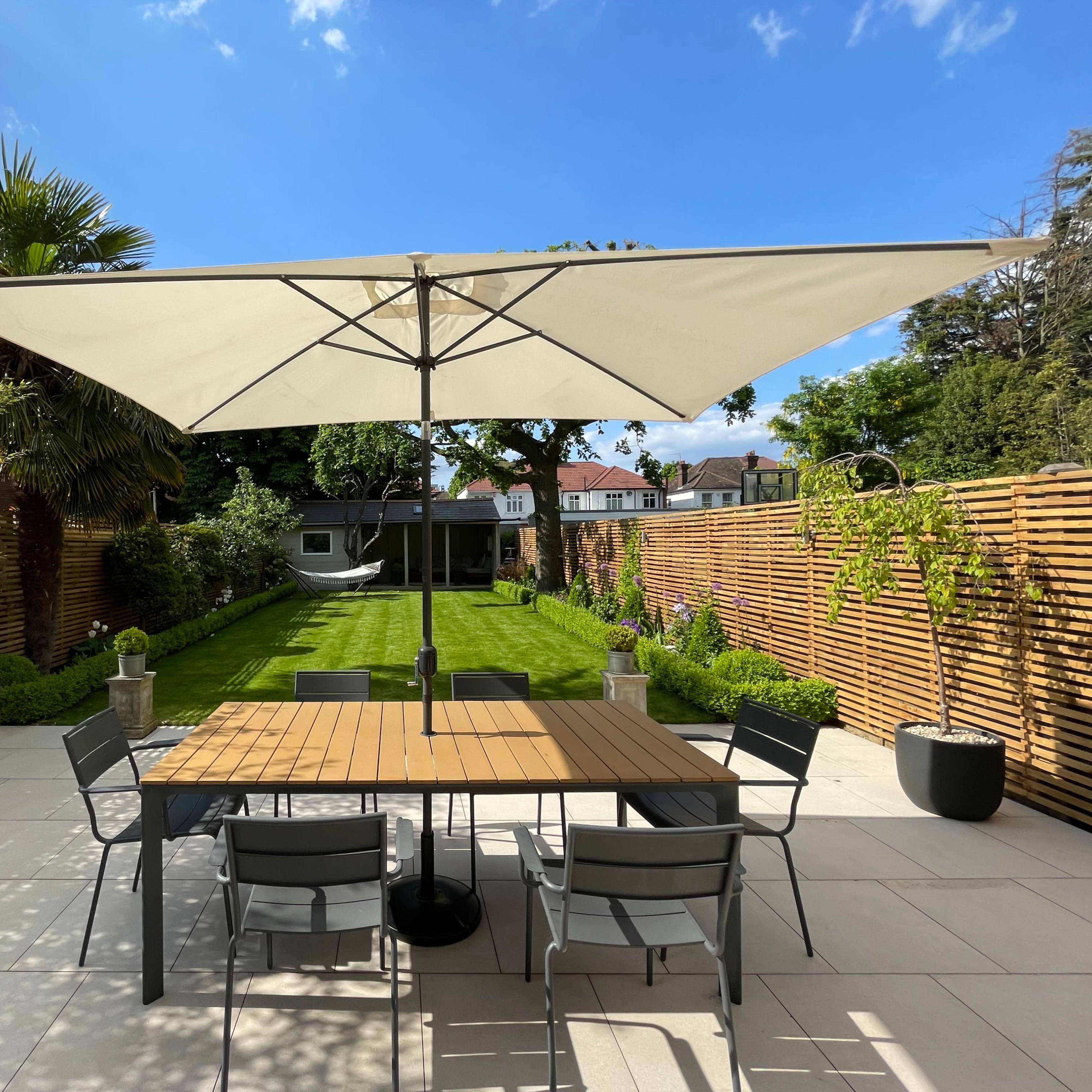 A large outdoor dining table is set beneath a spacious umbrella in a well-maintained garden with a lawn and wooden fencing.