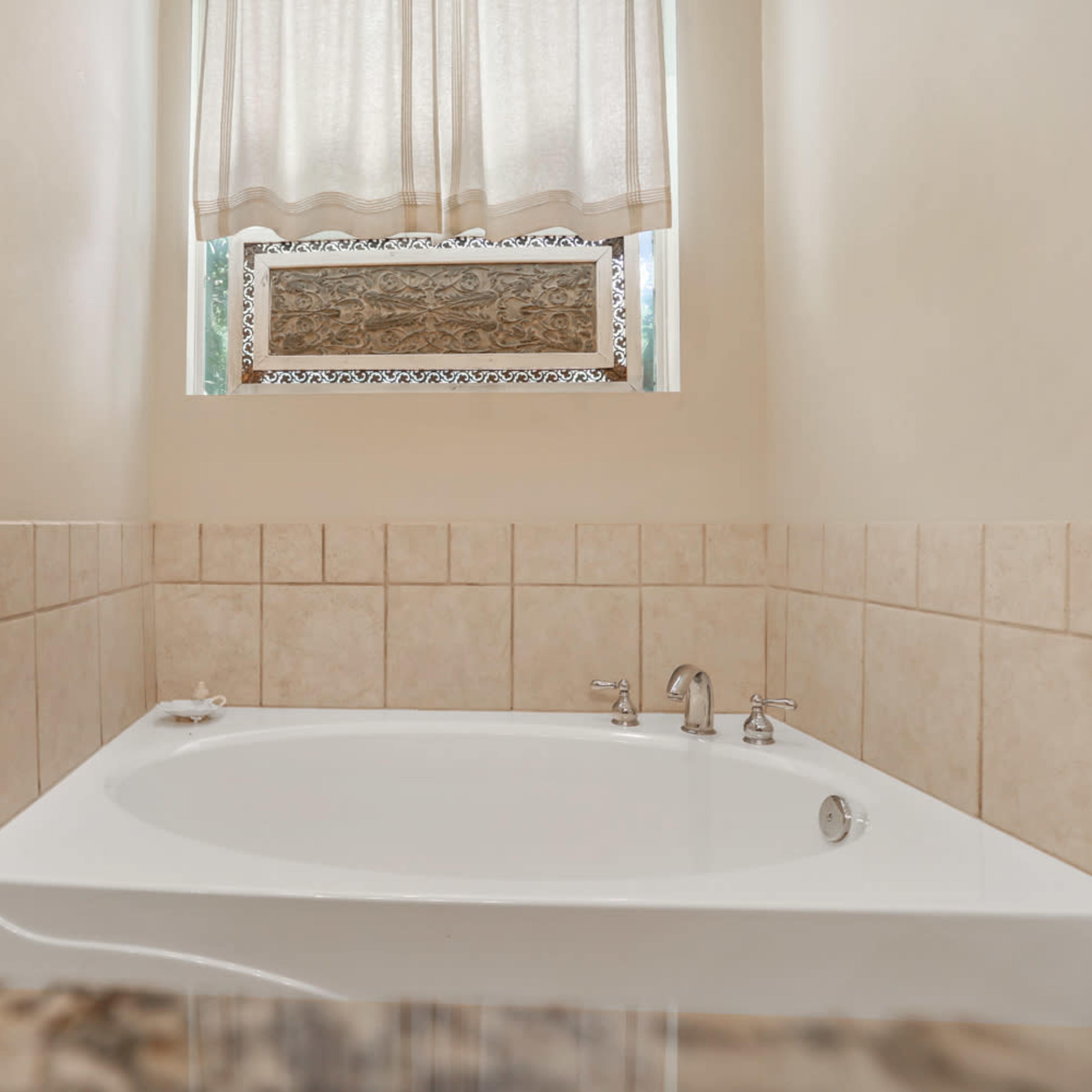 A white, oval bathtub is positioned next to a window with sheer curtains, surrounded by beige tiled walls.