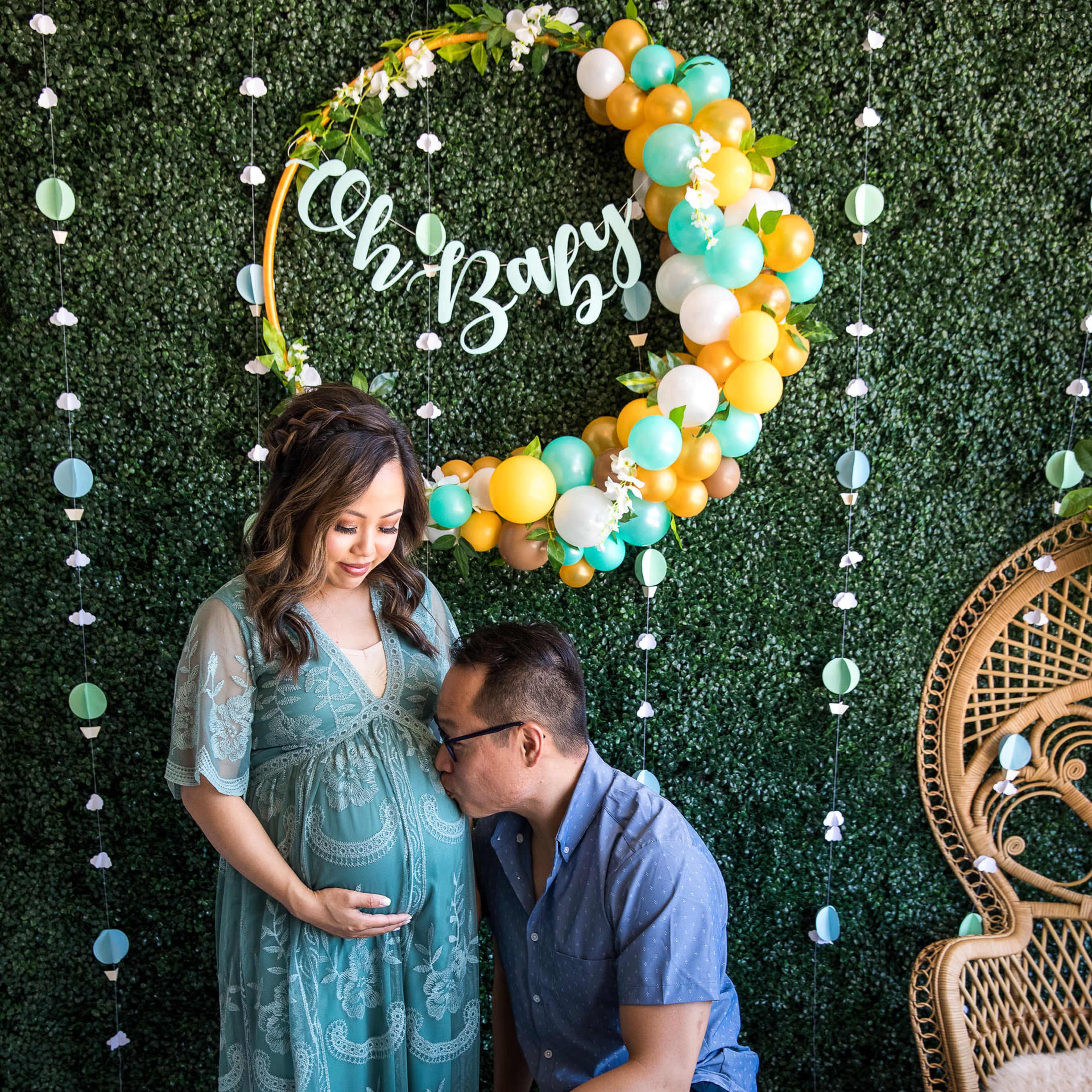 A woman in a teal dress stands in front of a decorative green backdrop, while a man kneels beside her, kissing her baby bump.