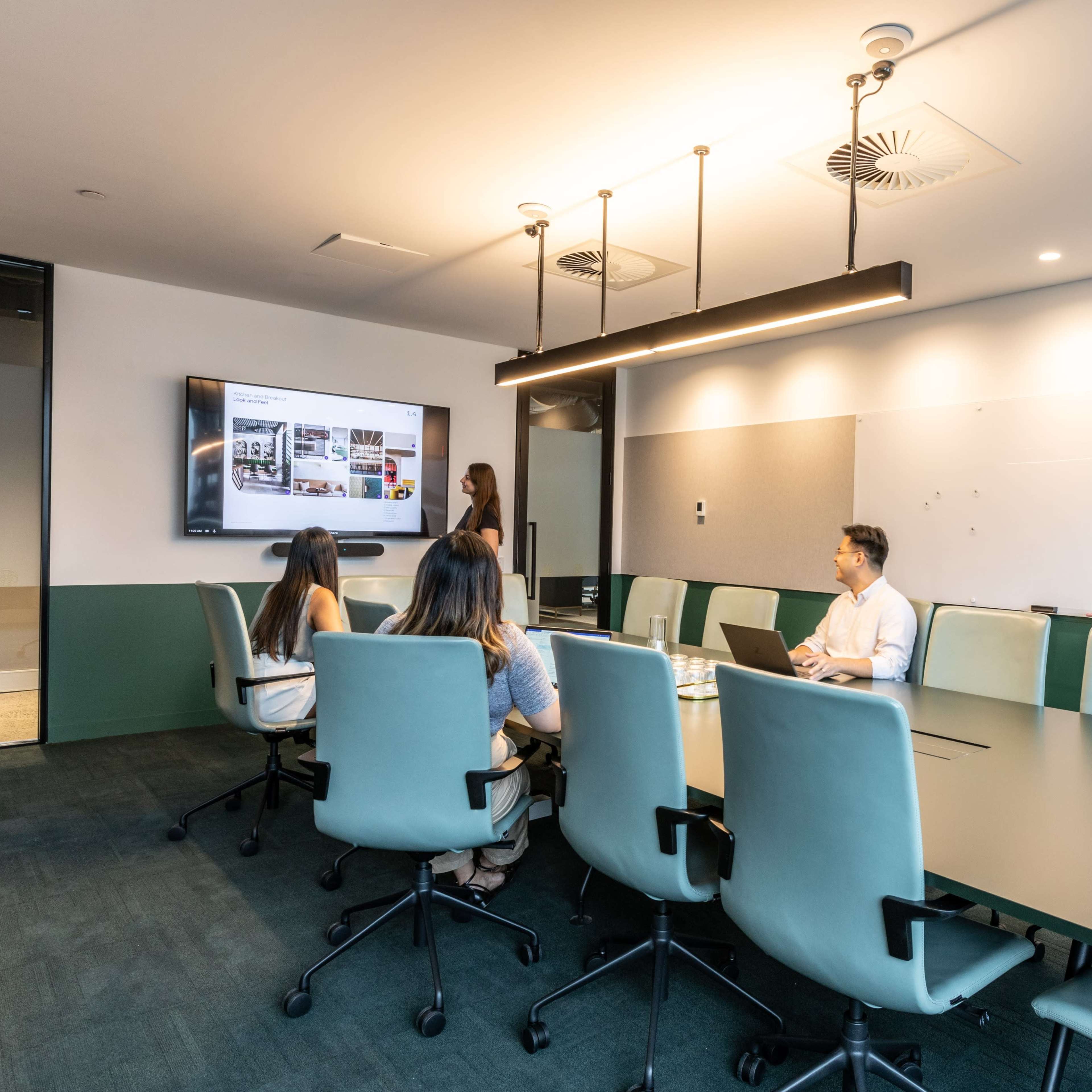 A group of five people sits around a large conference table in a modern meeting room, with a screen displaying a presentation.