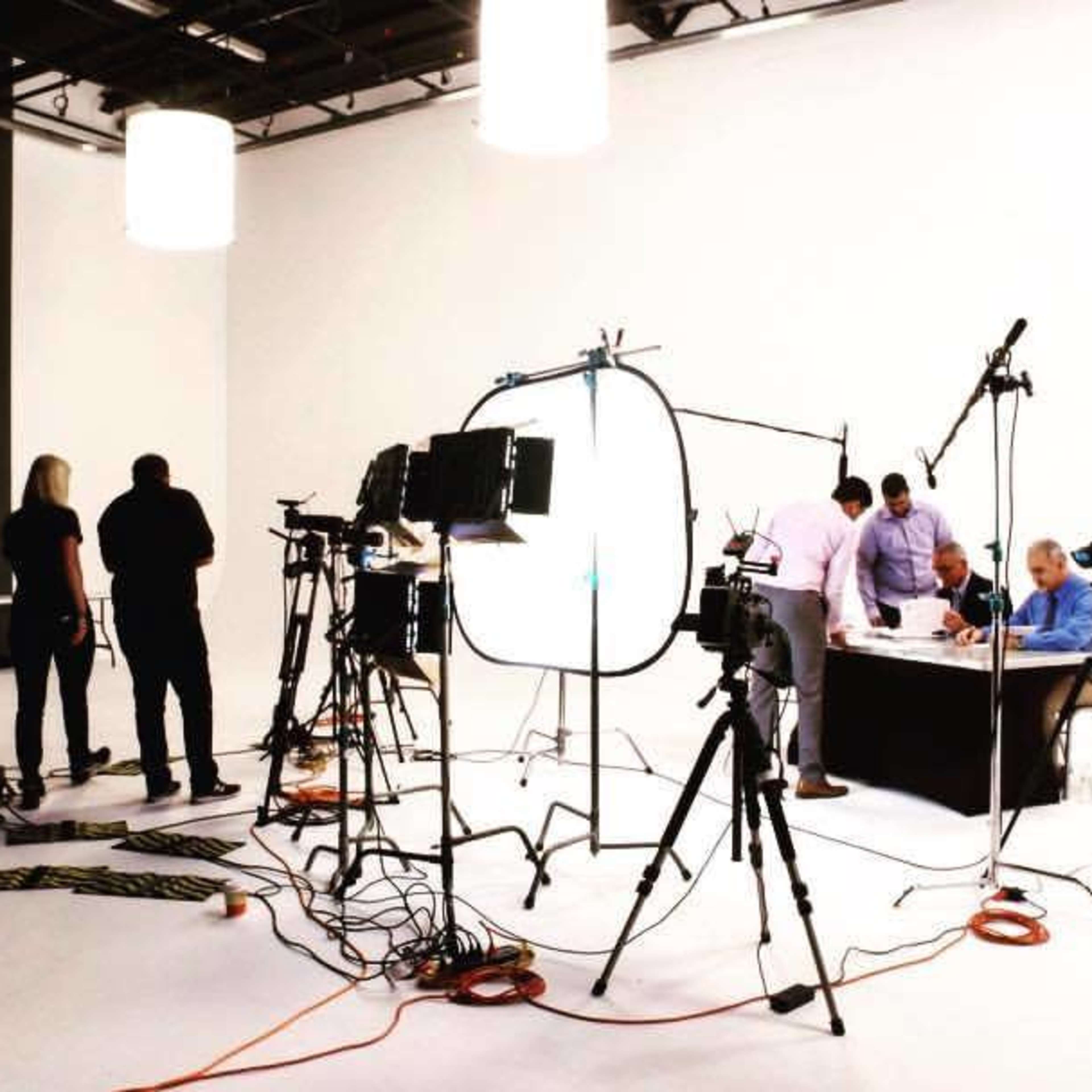 A film crew sets up equipment in a bright studio featuring a table where people are gathered for a recording.