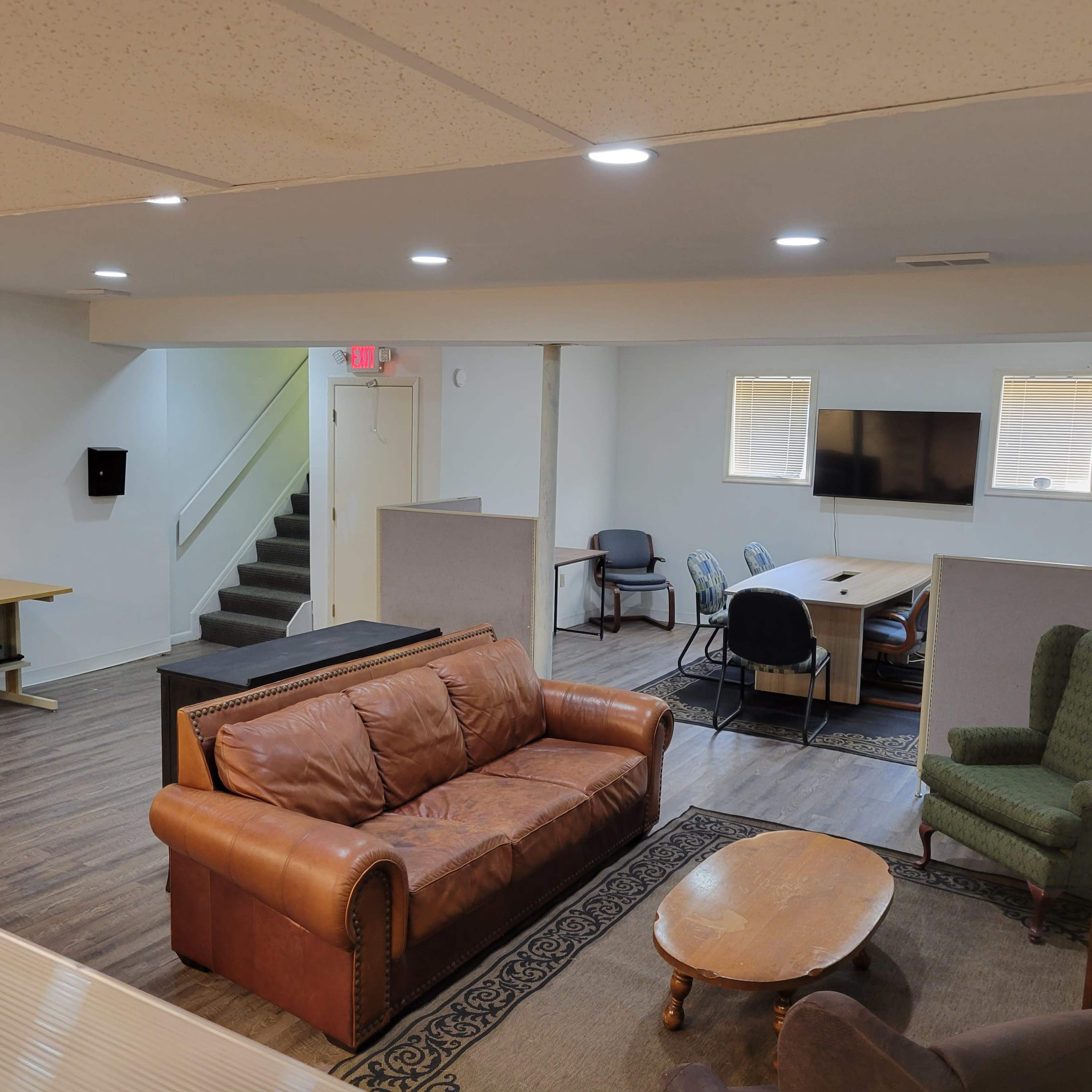 A spacious, multi-purpose room with a brown leather sofa, a wooden coffee table, and various seating arrangements, including a desk and an area with a television.