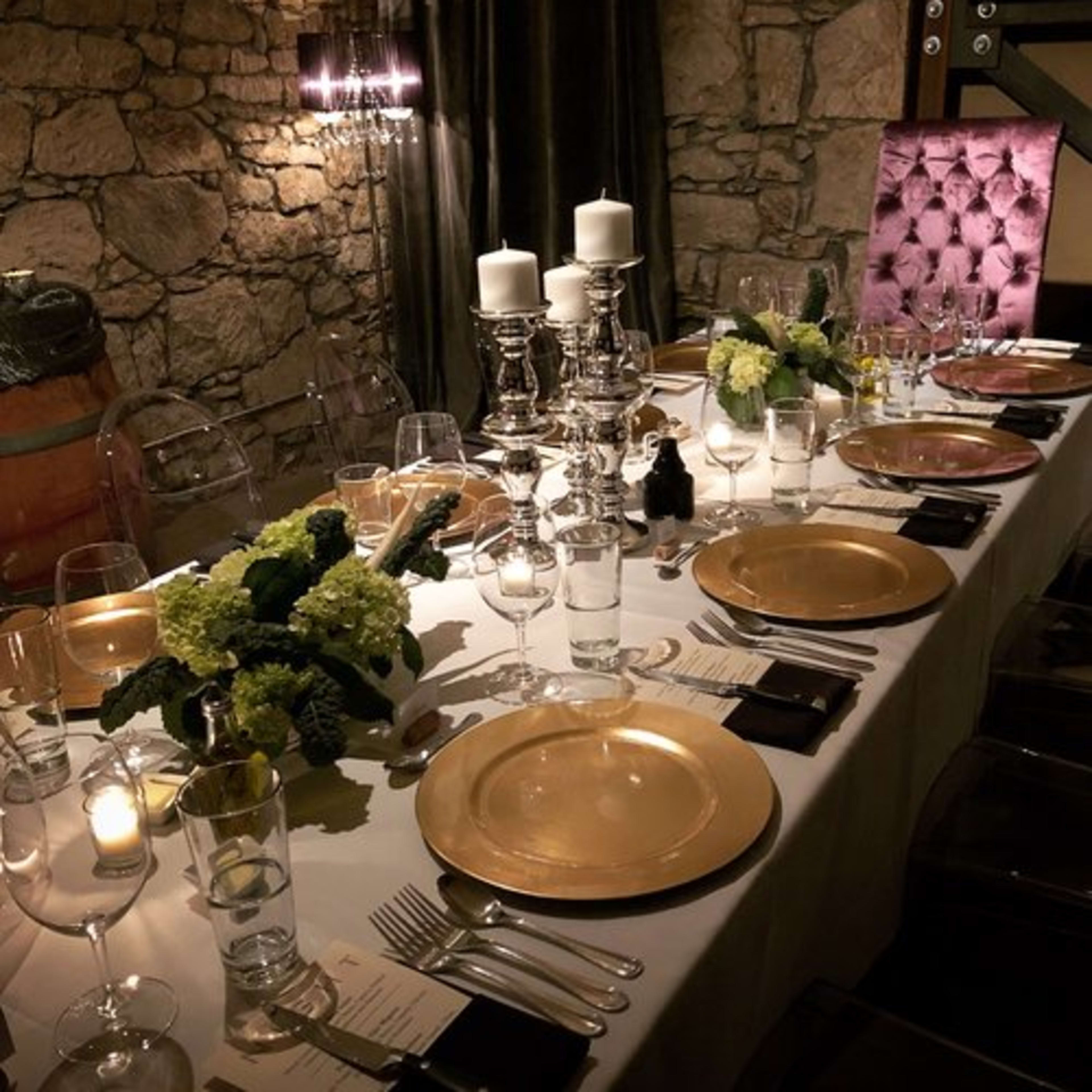 A elegantly set dining table features gold charger plates, glassware, candles, and floral arrangements against a stone wall backdrop.