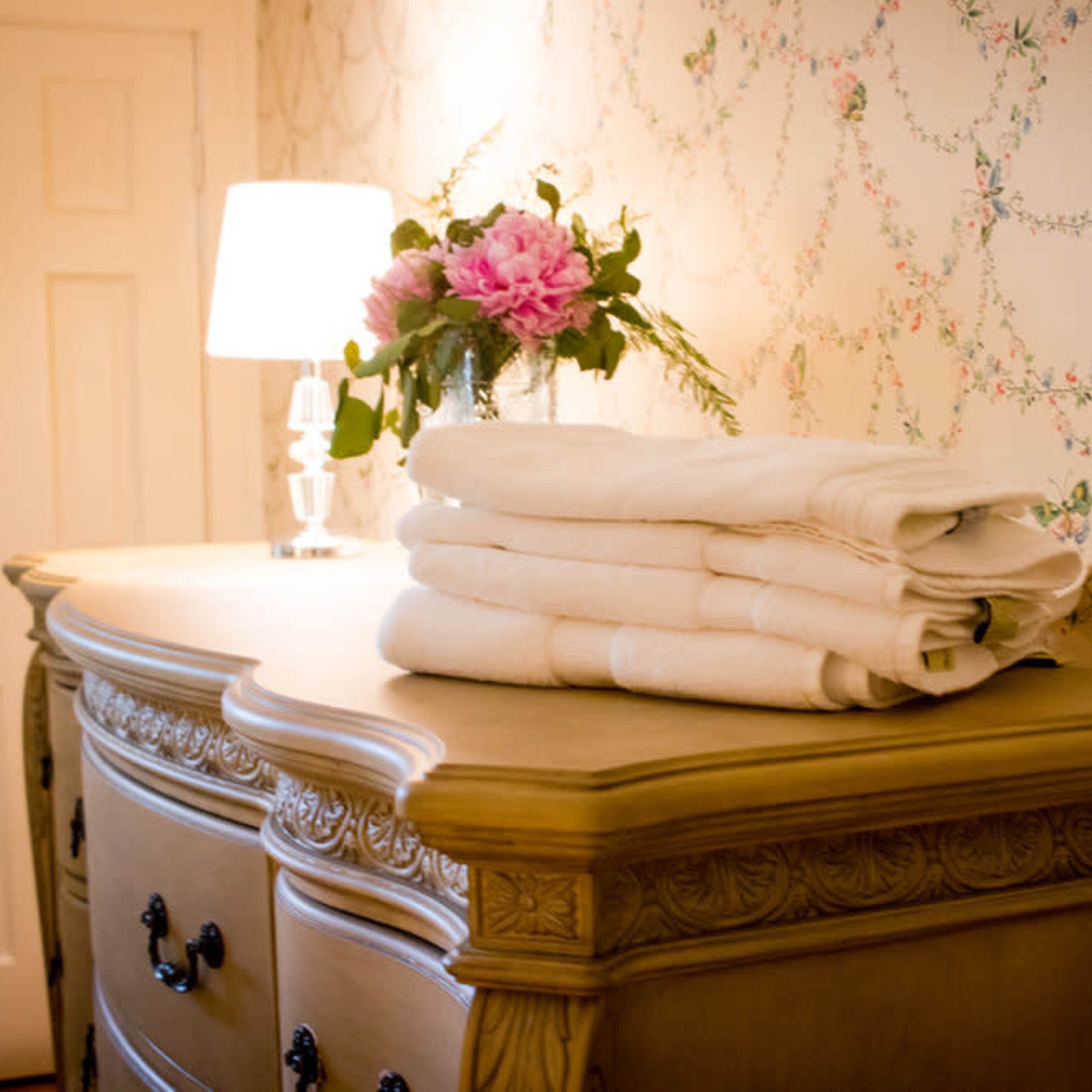 A stack of neatly folded white towels sits on top of a decorative wooden dresser beneath a lamp and a floral arrangement against a patterned wallpaper.