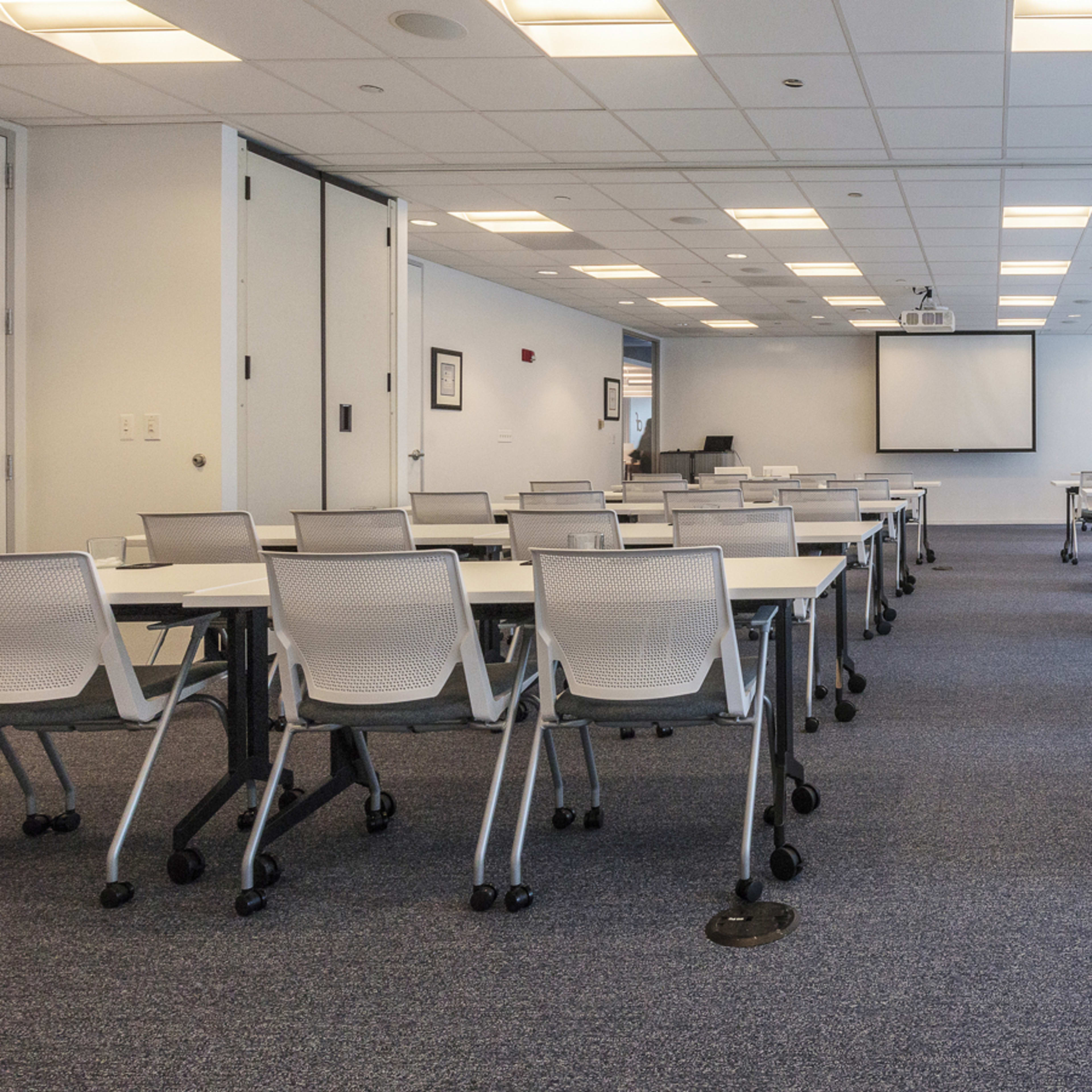 A large conference room filled with multiple rows of white tables and rolling chairs, facing a blank projector screen at the front.