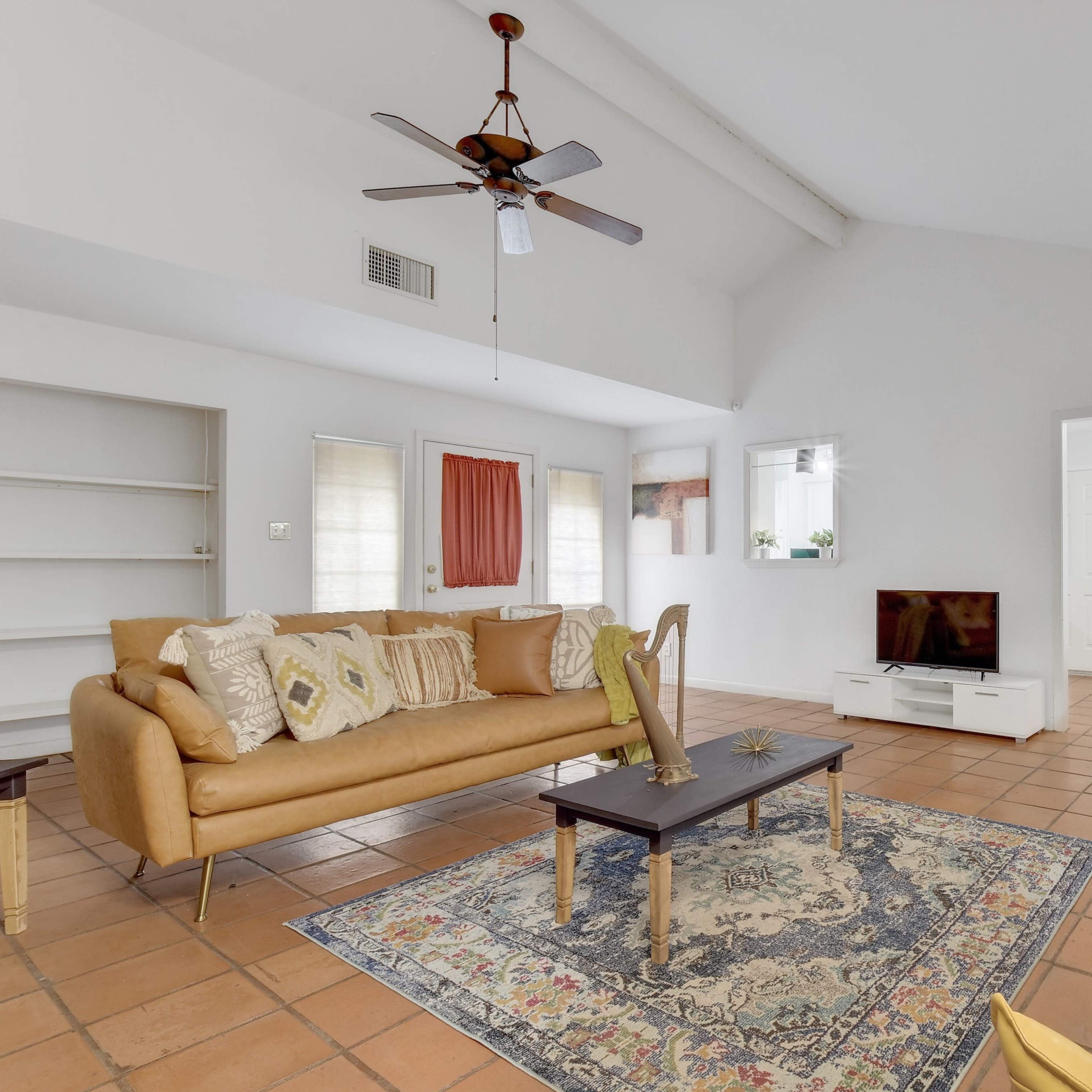 A living room features a tan sofa, a black coffee table on a patterned rug, and a television against a light-colored wall, with a dining area visible in the background.