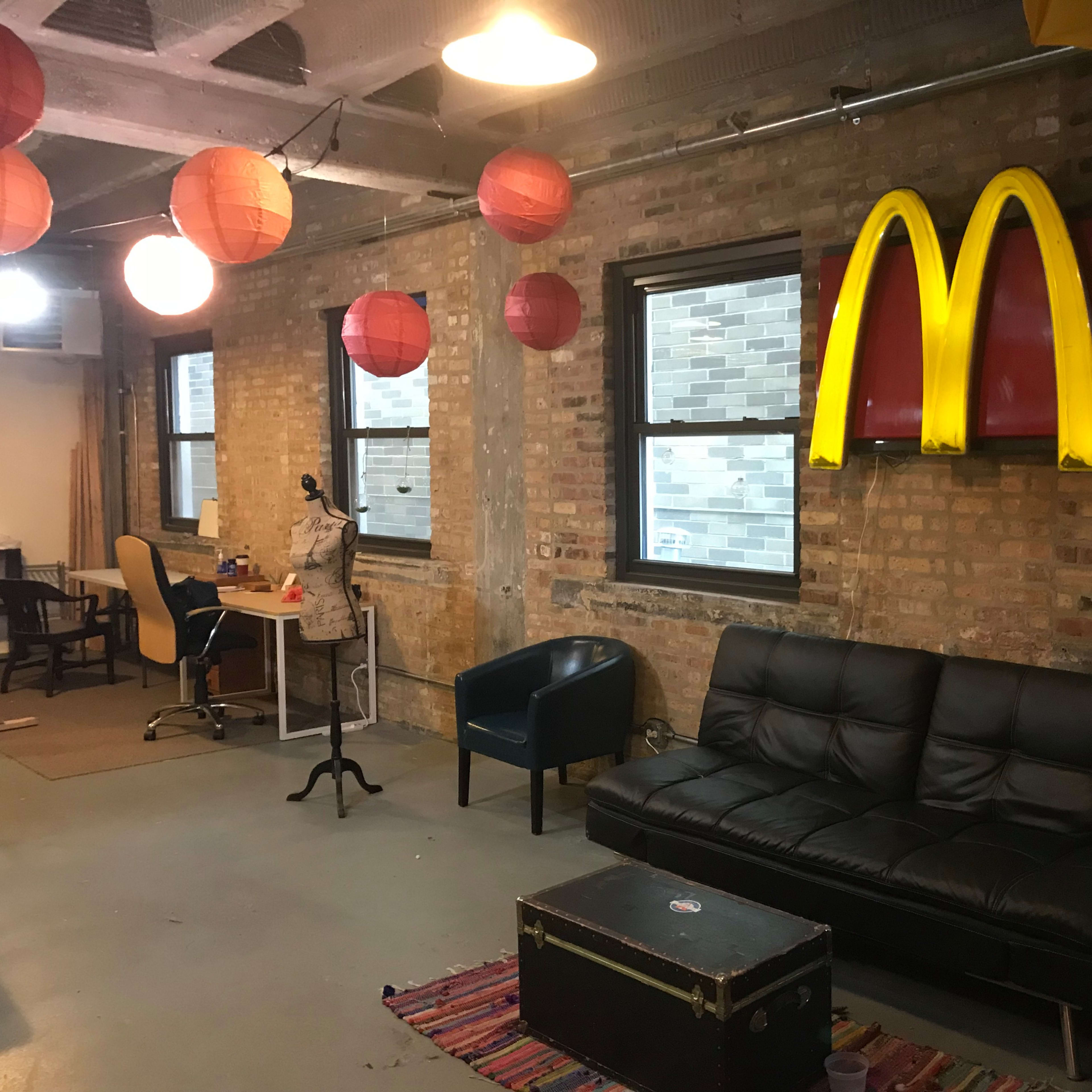 A casual office space featuring a black couch, a coffee table, a desk with a chair, and hanging red lanterns, with a large McDonald's logo displayed on the wall.