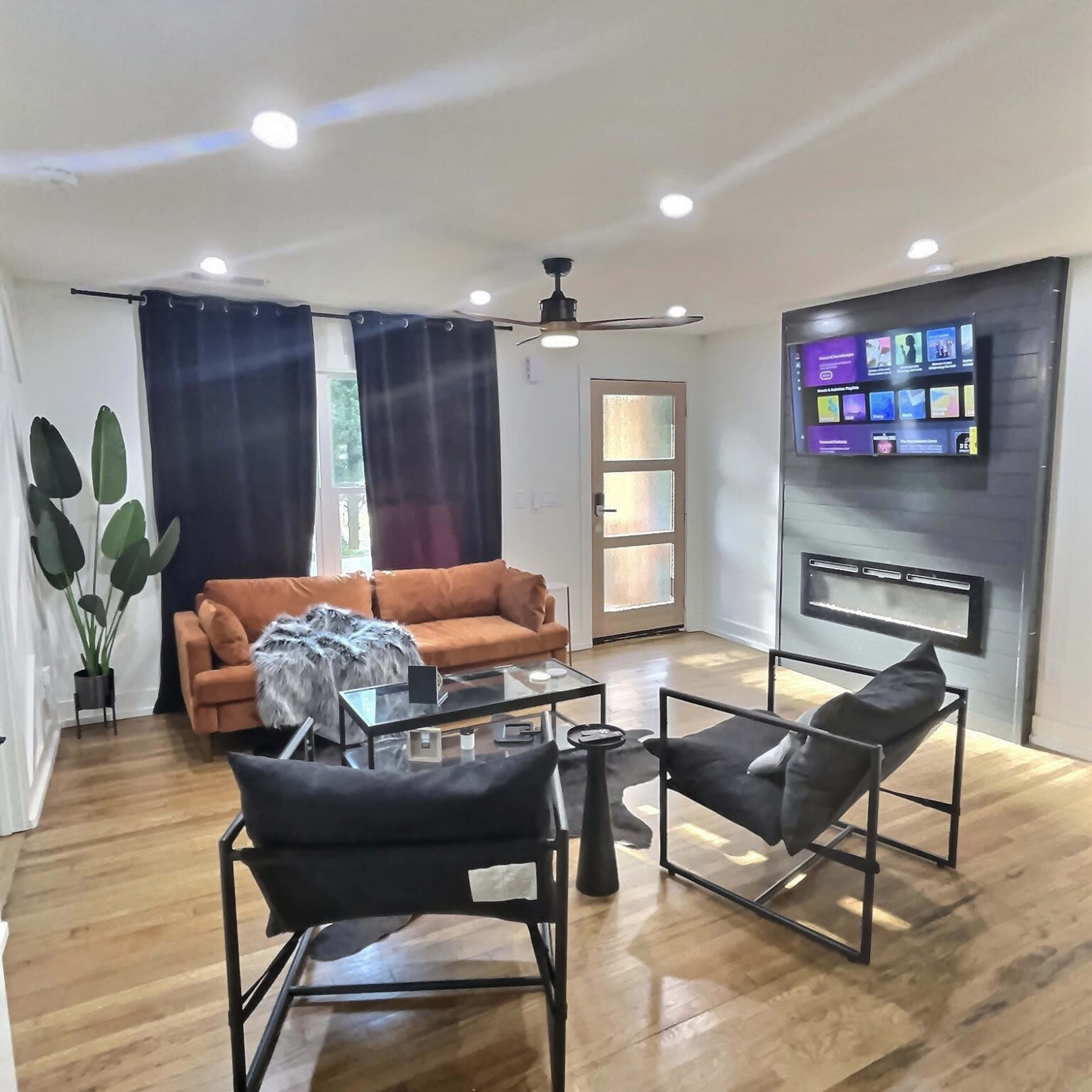 A modern living room with a brown couch, black chairs, a glass coffee table, and a mounted television above a fireplace.
