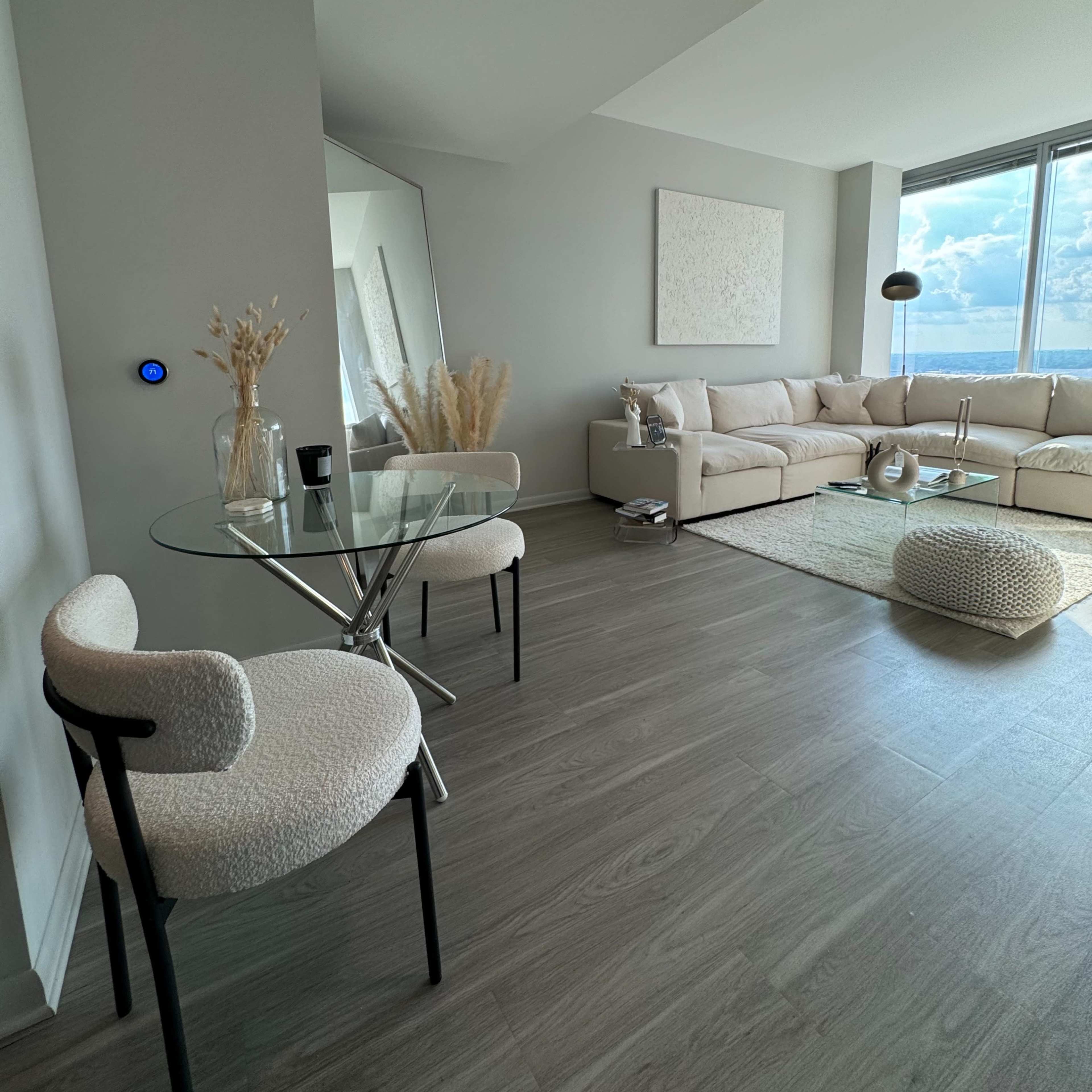 A modern living room features a beige sectional sofa, a glass coffee table, and two chairs, with large windows showcasing a view outside.
