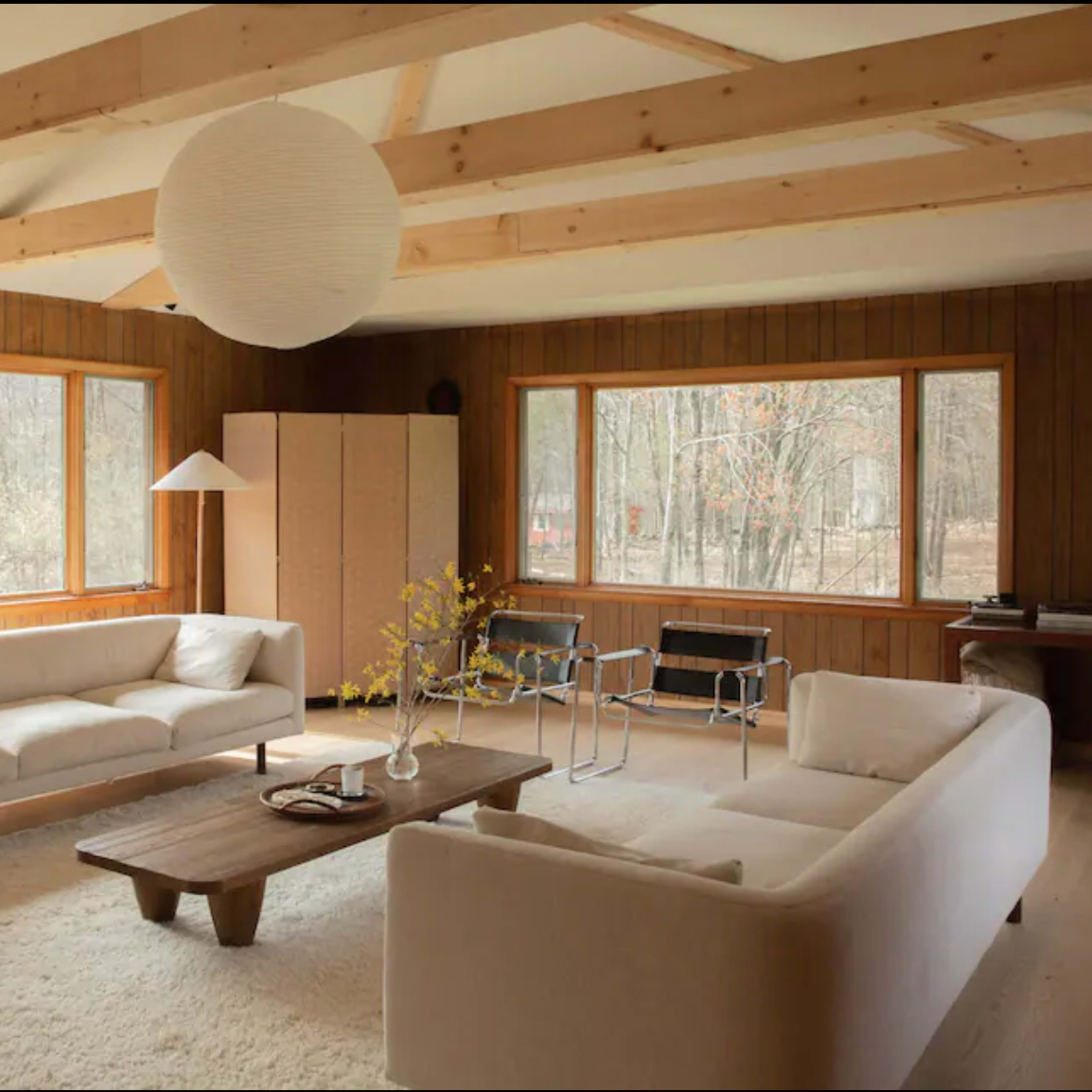 A living room features two white sofas, a wooden table, and large windows overlooking a wooded area, with minimalist furnishings and a paper lantern ceiling light.