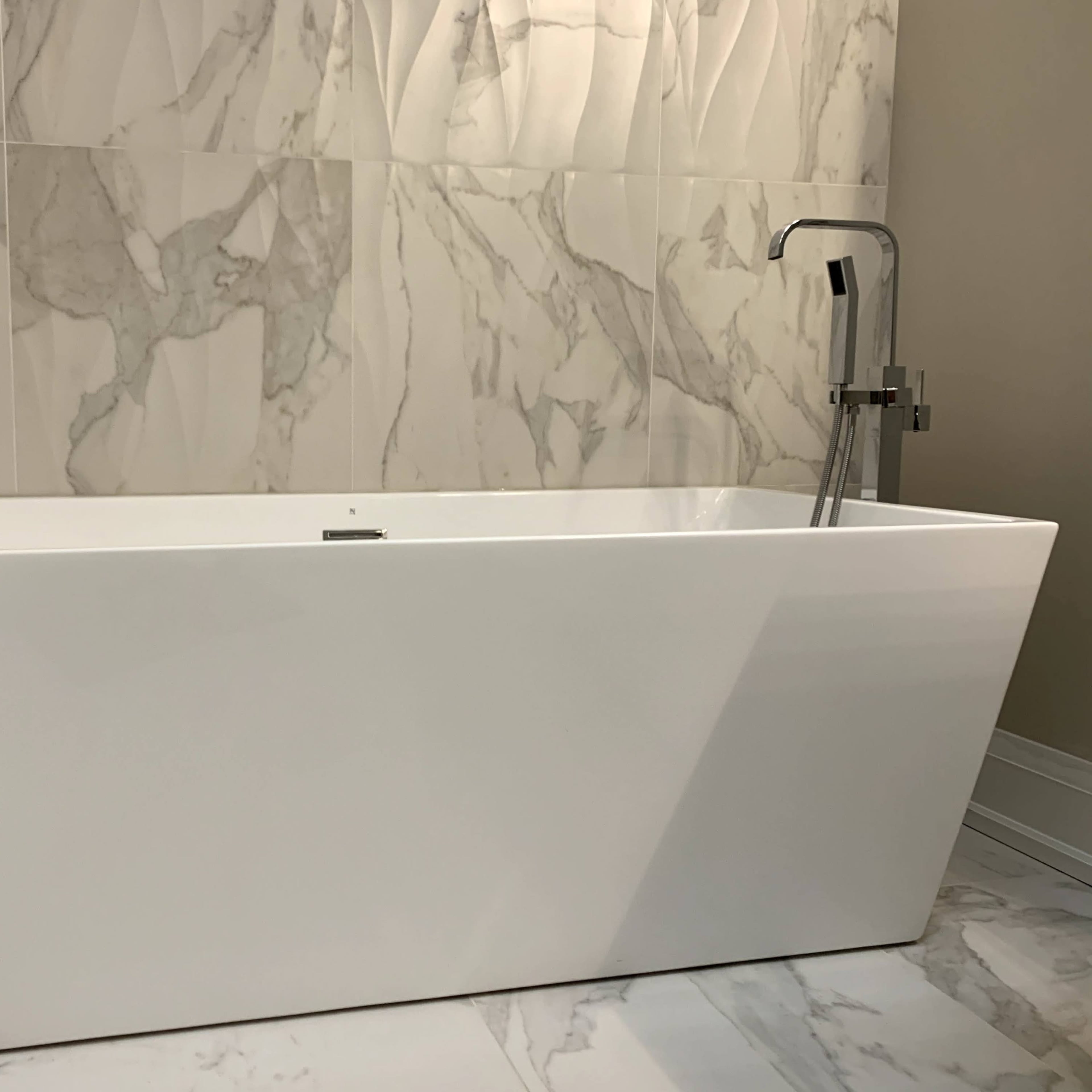 A modern, white freestanding bathtub in a bathroom with marble-patterned walls and flooring.