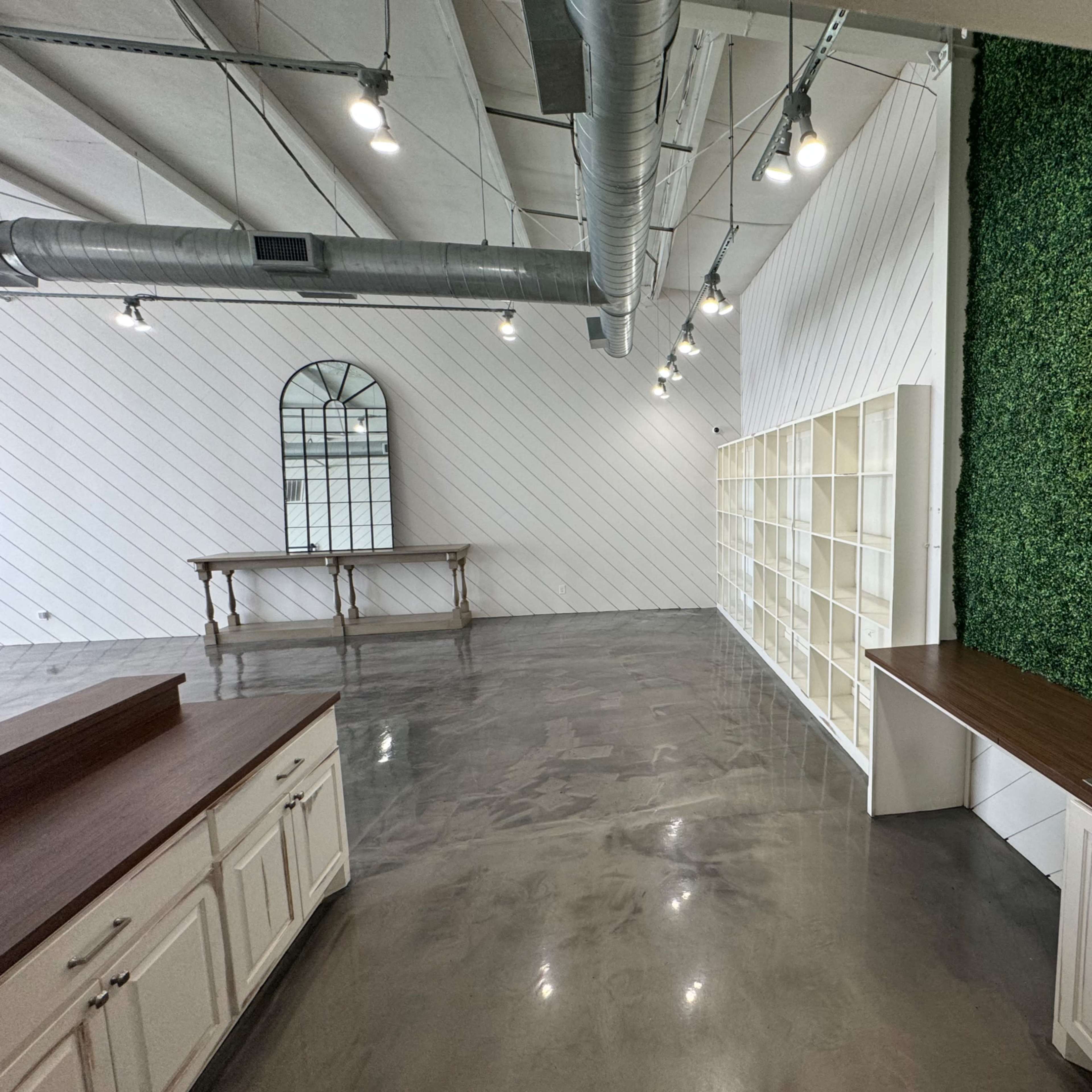 A spacious room with a concrete floor, a green vertical wall, shelving units, a sink, and overhead lights.