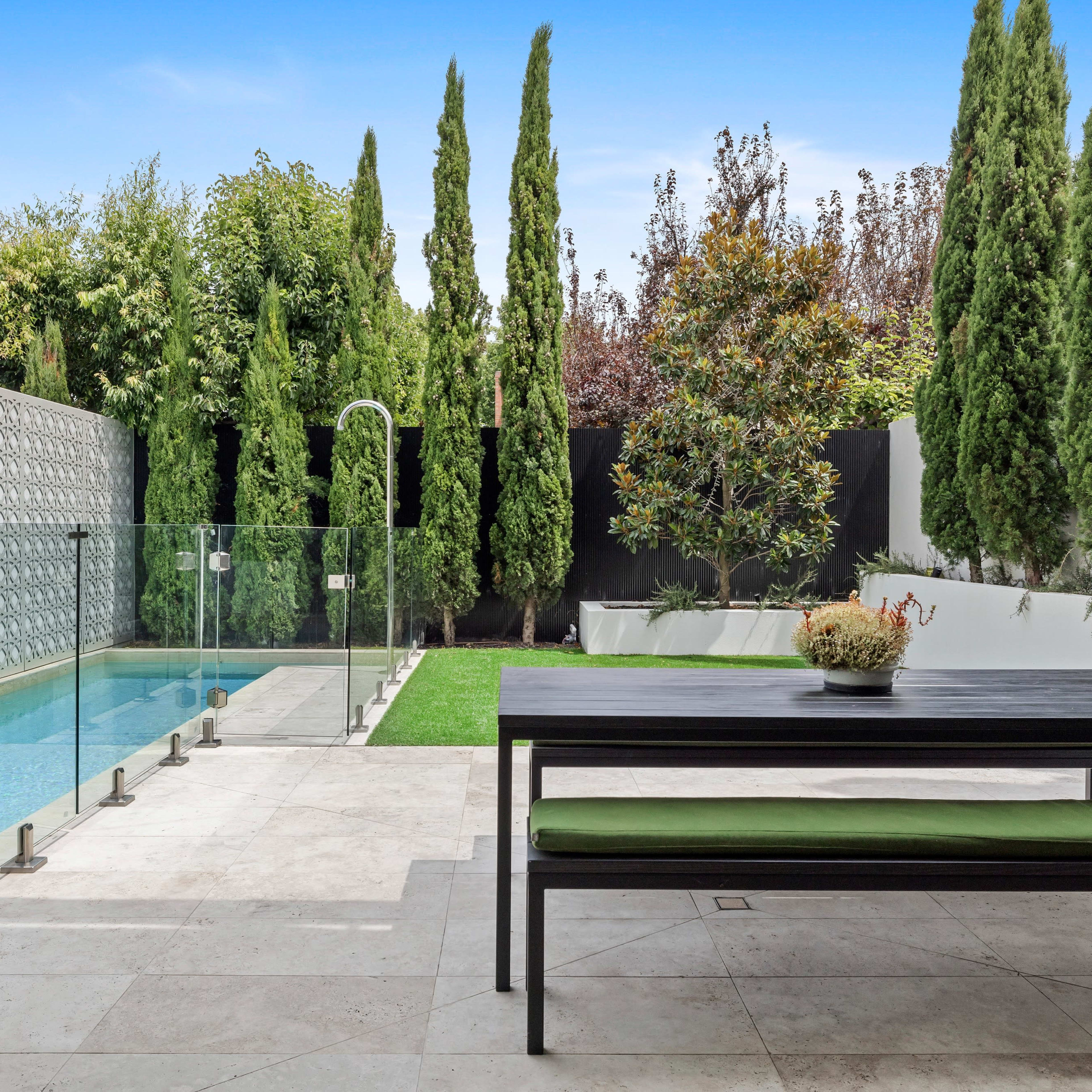 A modern outdoor area featuring a swimming pool, a glass barrier, and a wooden dining table surrounded by tall evergreen trees.