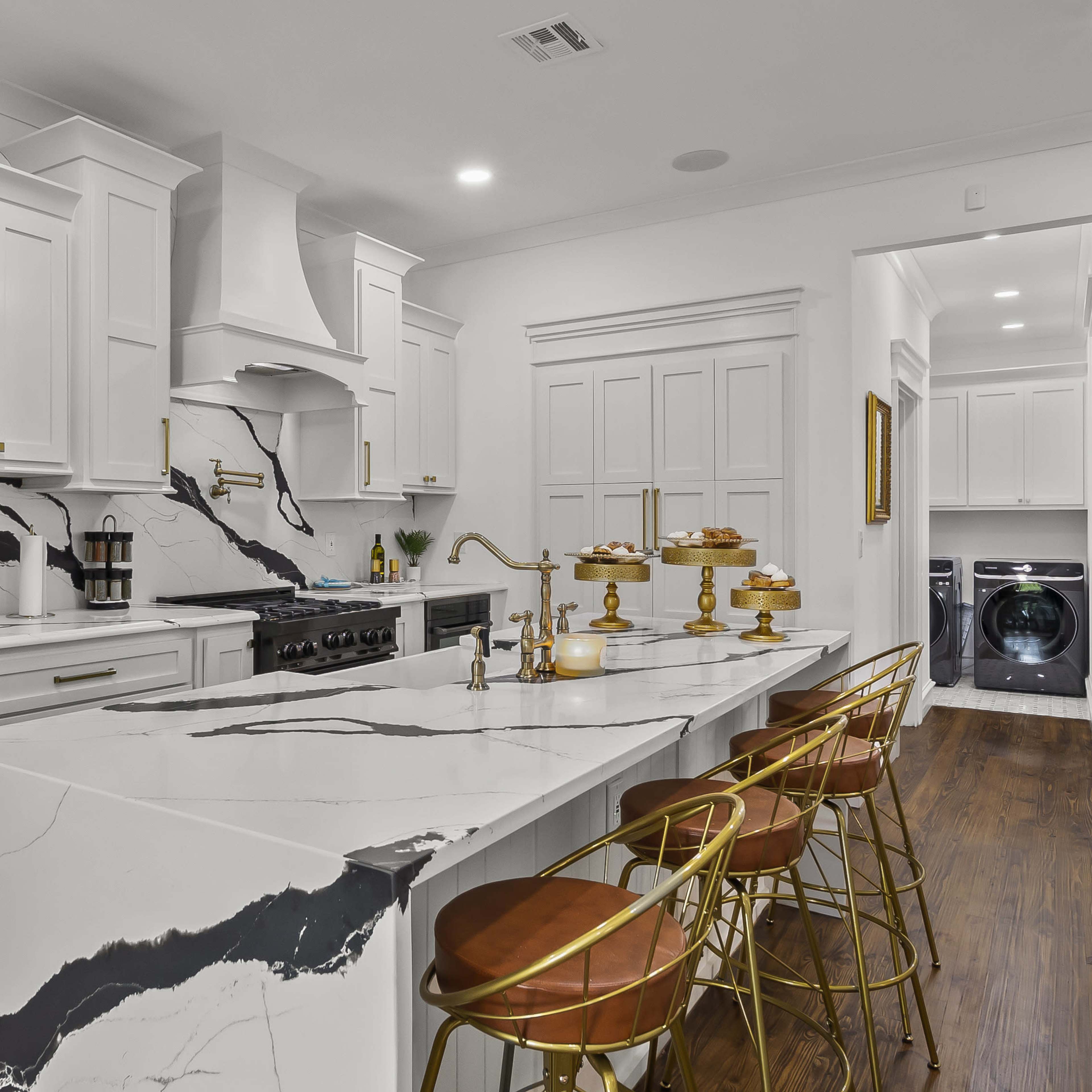 A modern kitchen with white cabinetry, a large marble island with gold bar stools, and stainless steel appliances, including a refrigerator and a range.