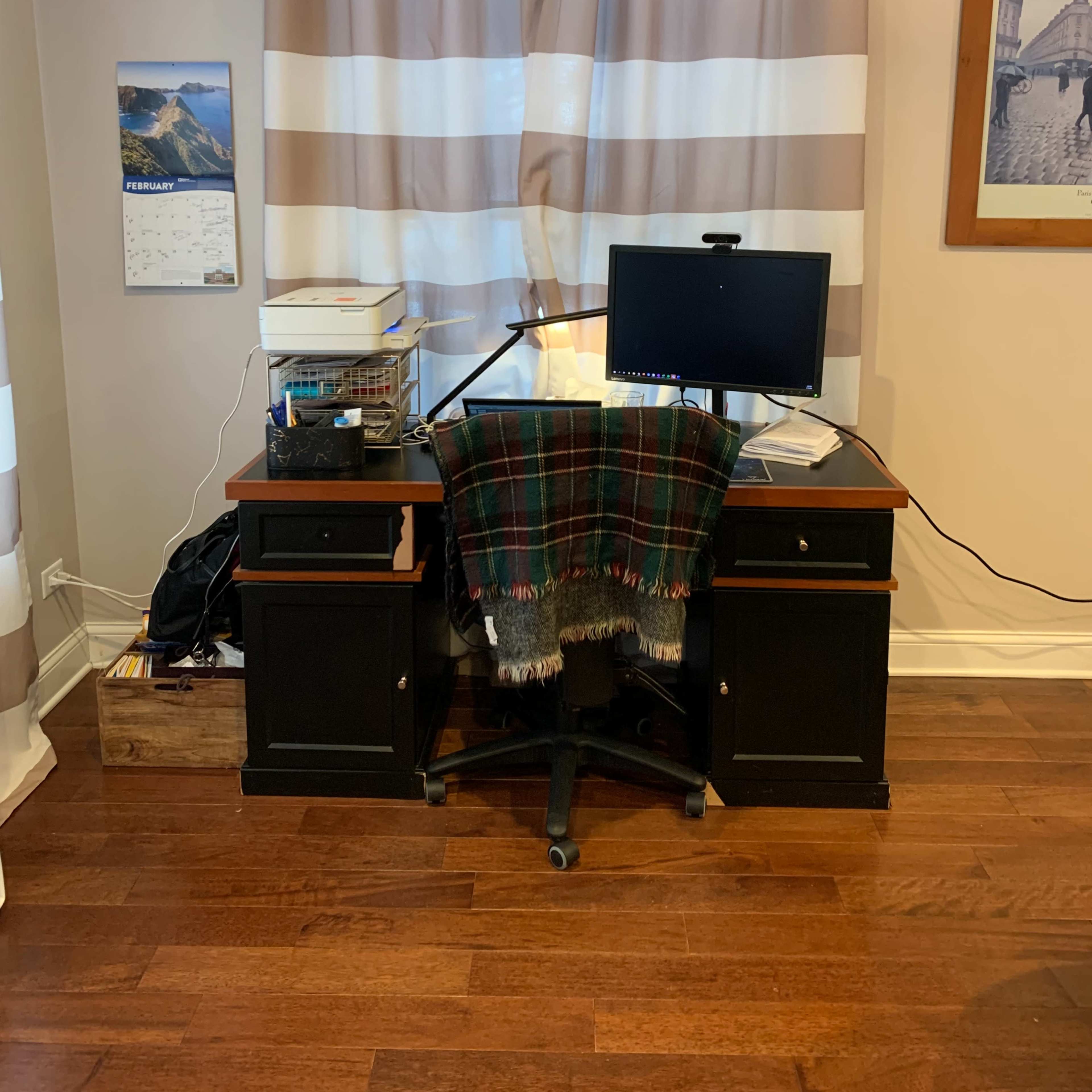 A home office setup features a black desk with a computer monitor, a chair, and a window adorned with striped curtains.